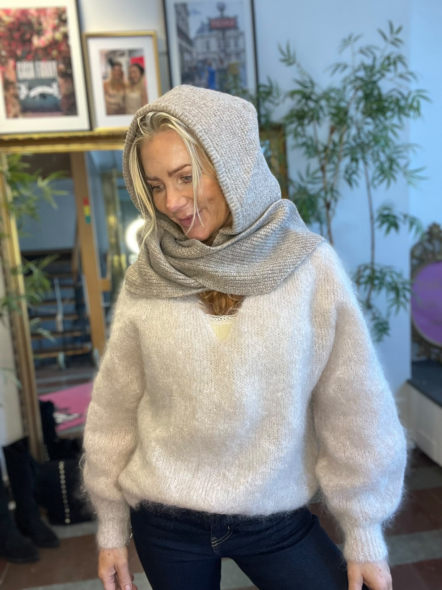 Kvinna med blondt hår bär en grå stickad sjal som huva och halsduk och ler nedåt. Hon står inomhus i ett rum med växter och inramade foton i bakgrunden.