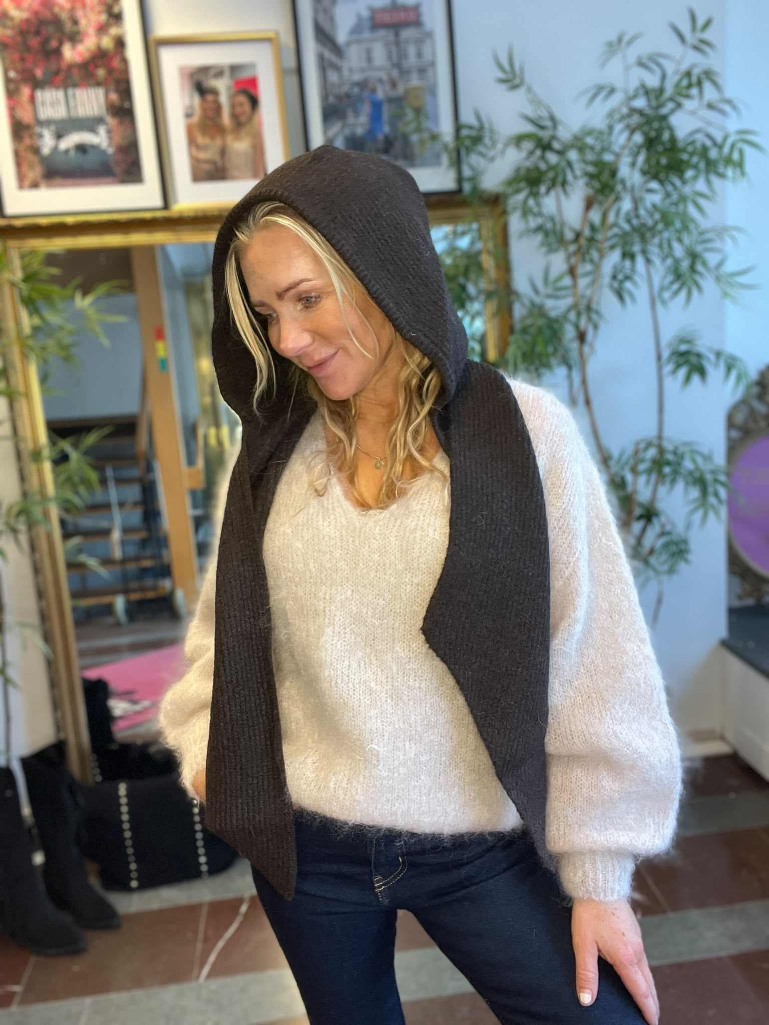 Blond kvinna står inomhus och tittar nedåt med ett svagt leende, iklädd vit stickad tröja och mörk huva/sjal över huvudet. I bakgrunden syns inramade tavlor och en hög grön växt.