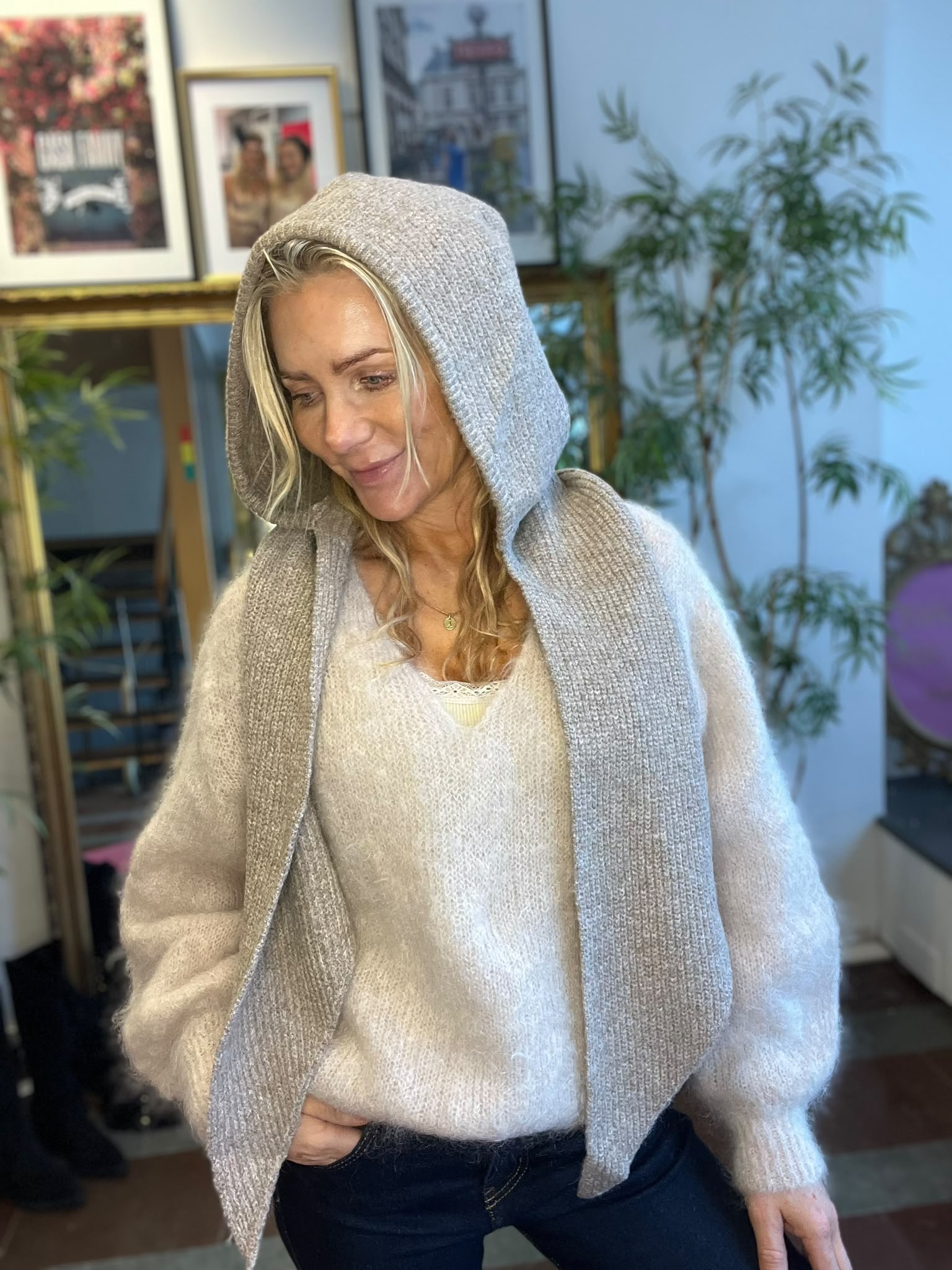 Blond kvinna i ljus, fluffig tröja och grå stickad sjal med huva tittar ner och ler svagt. Hon står inomhus framför inramade foton och en stor grön växt, i mjukt dagsljus.