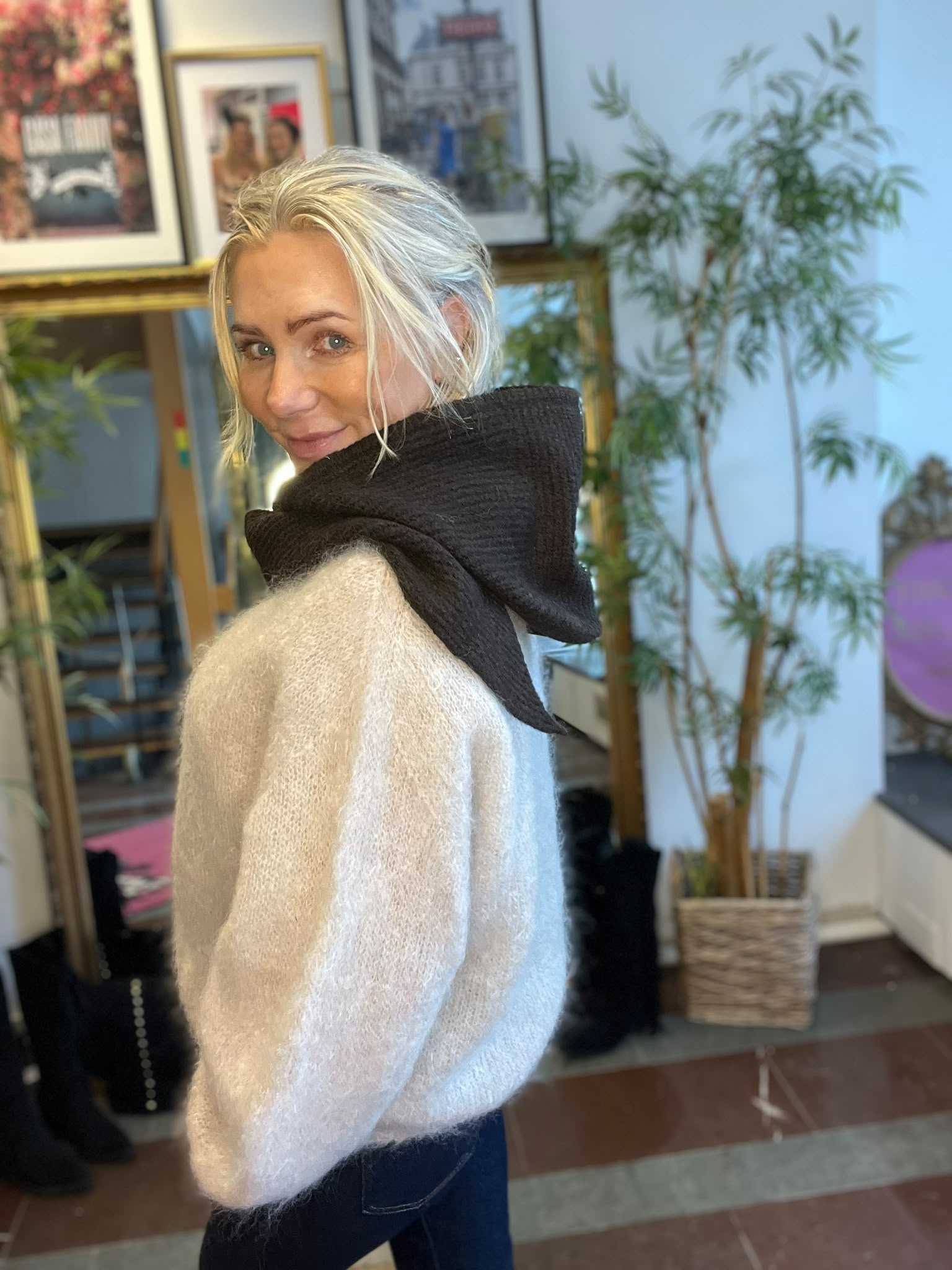 Blond person i ljus stickad tröja och stor svart halsduk vänder sig om och ler mot kameran. Står inomhus framför en spegel med inramade bilder på väggen och en stor krukväxt i bakgrunden.