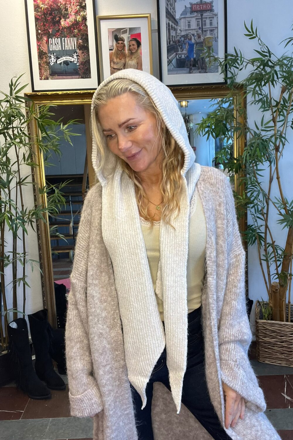 Blond kvinna i beige kofta, vit halsduk och uppdragen luva lutar sig lätt fram och ler. Hon står bland gröna krukväxter framför spegel och inramade foton; ett foto har texten “CASA FANNY”.