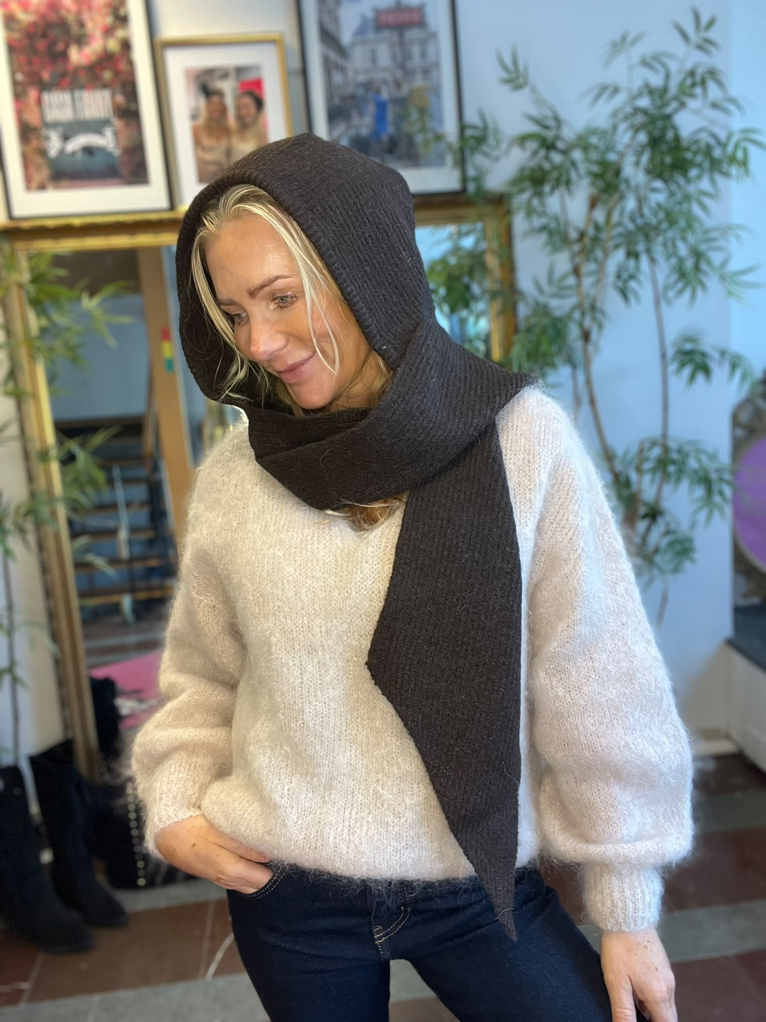 Blond kvinna står inomhus och tittar ned med ett litet leende, iklädd vit stickad tröja och mörk stickad sjal dragen som huva och lindad runt halsen. I bakgrunden syns växter och inramade affischer.