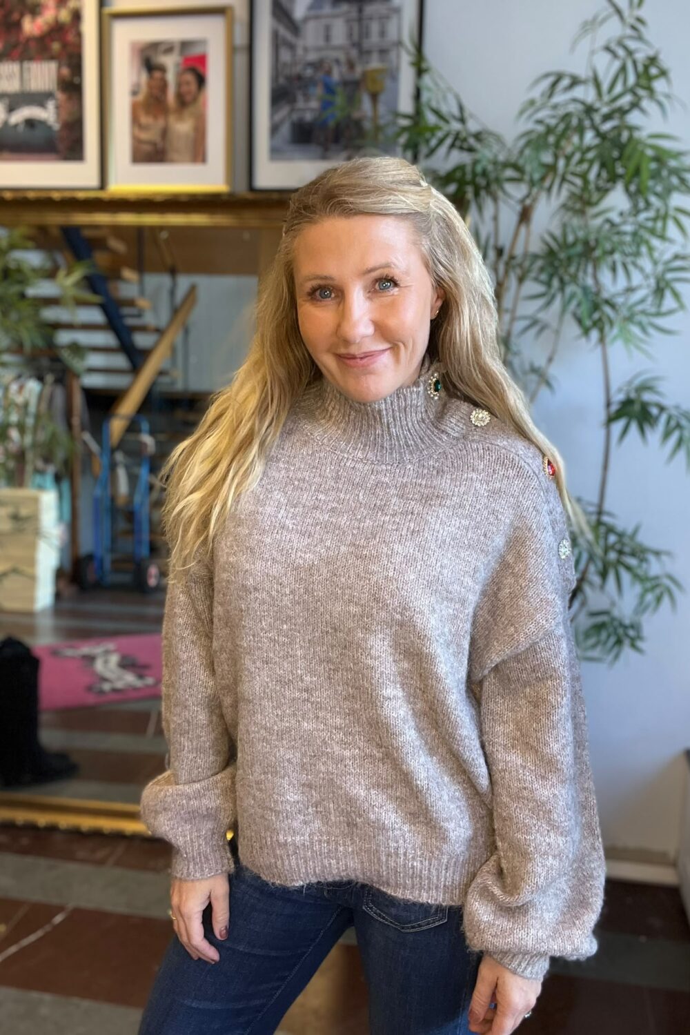 Leende kvinna med långt blont hår poserar inomhus i en ljusgrå stickad polotröja med dekorativa knappar och jeans. I bakgrunden syns gröna krukväxter samt en spegel och inramade fotografier på väggen.