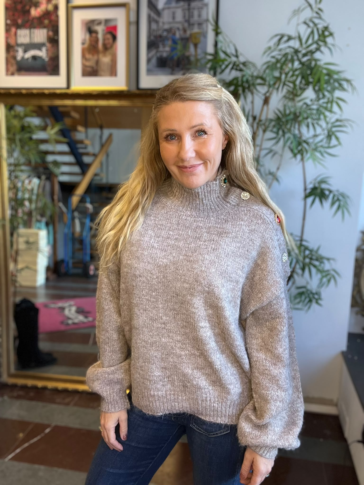 Leende kvinna med långt blont hår poserar inomhus i en ljusgrå stickad polotröja med dekorativa knappar och jeans. I bakgrunden syns gröna krukväxter samt en spegel och inramade fotografier på väggen.