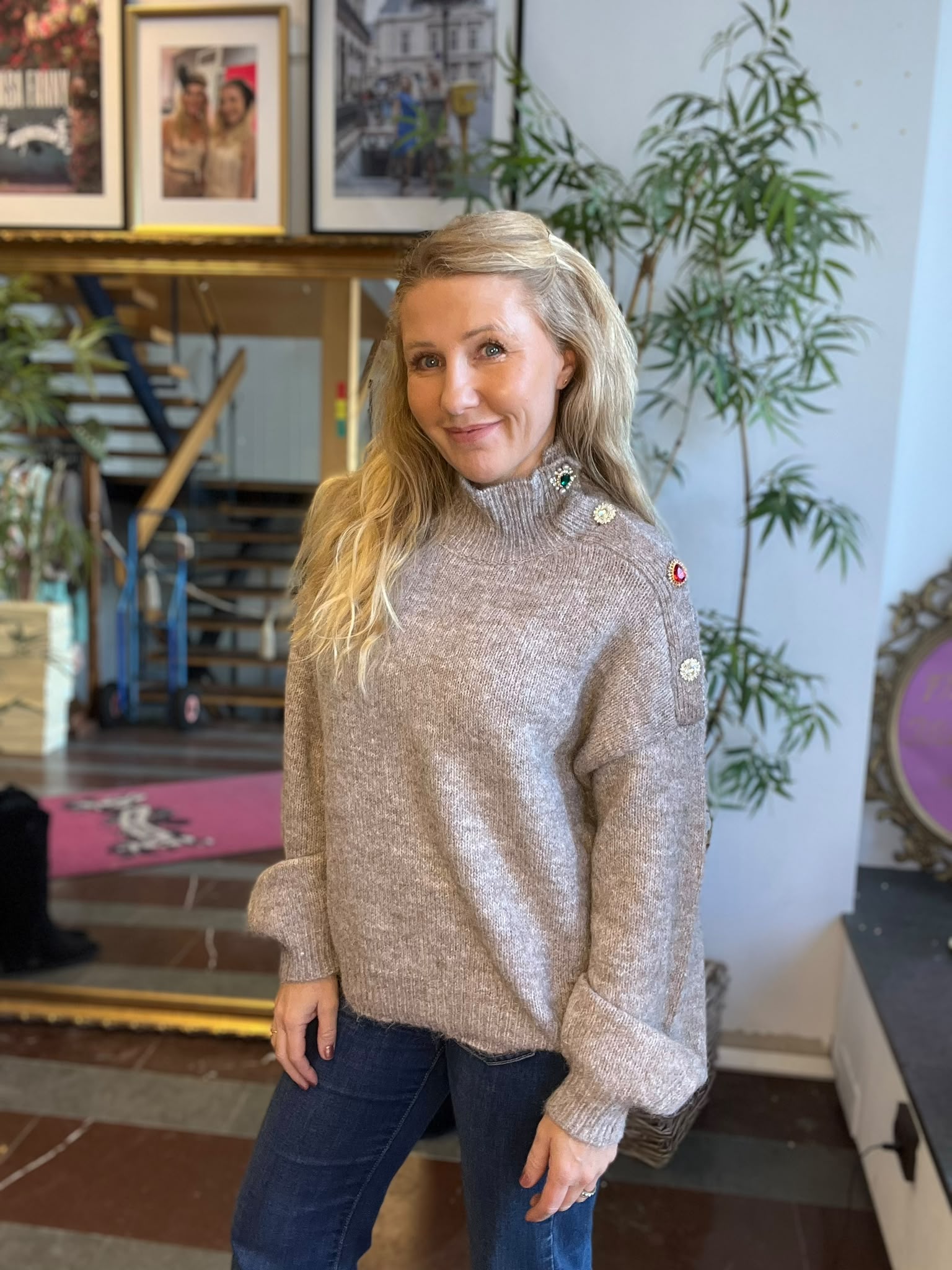 Leende kvinna med långt blont hår poserar och tittar mot kameran, klädd i grå stickad polotröja med dekorativa knappar och jeans. Hon står inomhus framför en trappa, inramade foton och en stor grön växt.