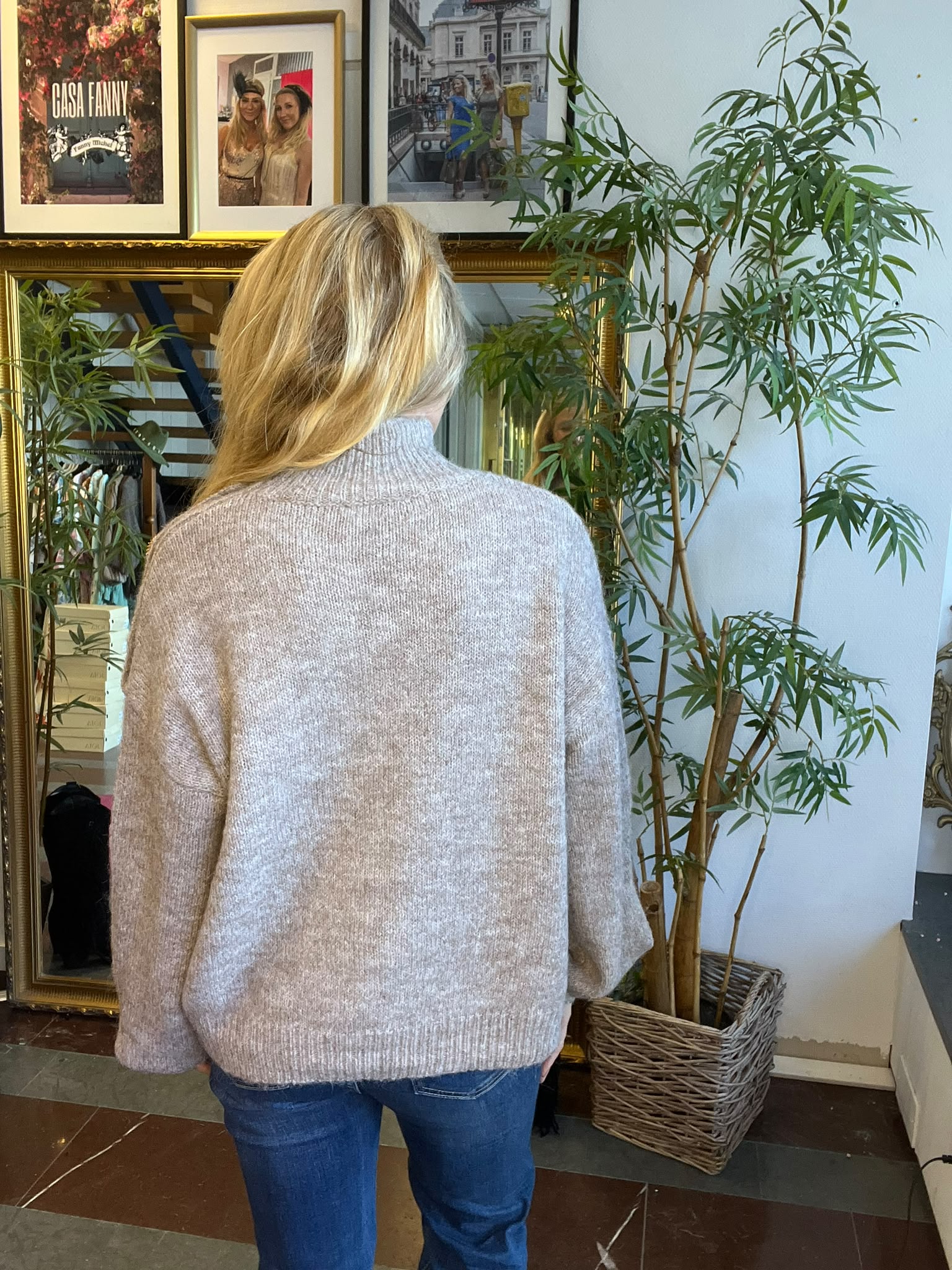 Kvinna med blondt hår står med ryggen mot kameran och tittar på inramade foton på en vägg. Bredvid henne står en hög grön krukväxt och en stor spegel. Text i en ram: ”CASA FANNY”.