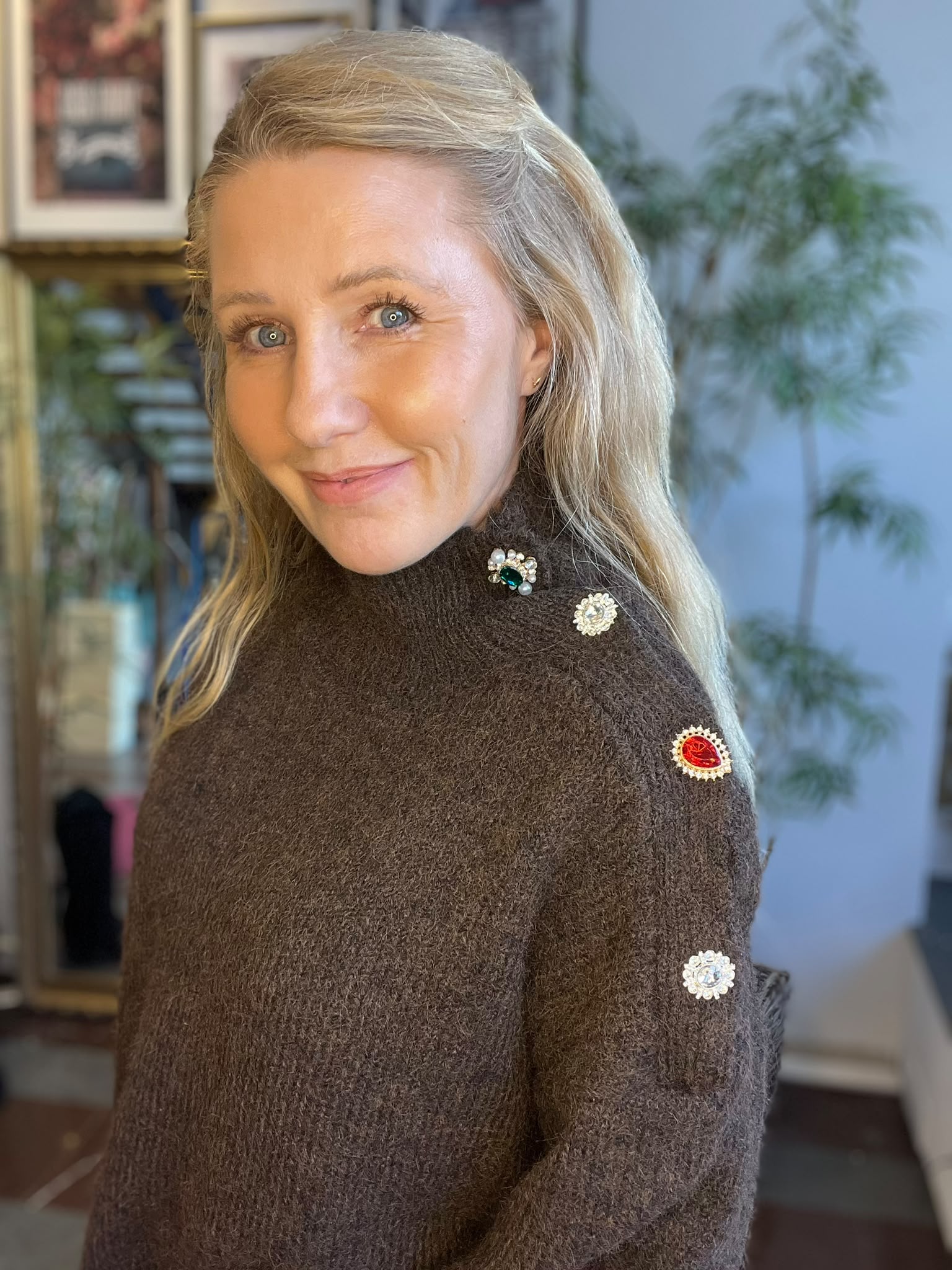 Leende kvinna med långt blont hår vänder sig mot kameran inomhus. Hon bär en brun stickad tröja dekorerad med gnistrande broscher i olika färger på axel och ärm, med suddig bokhylla och växt i bakgrunden.