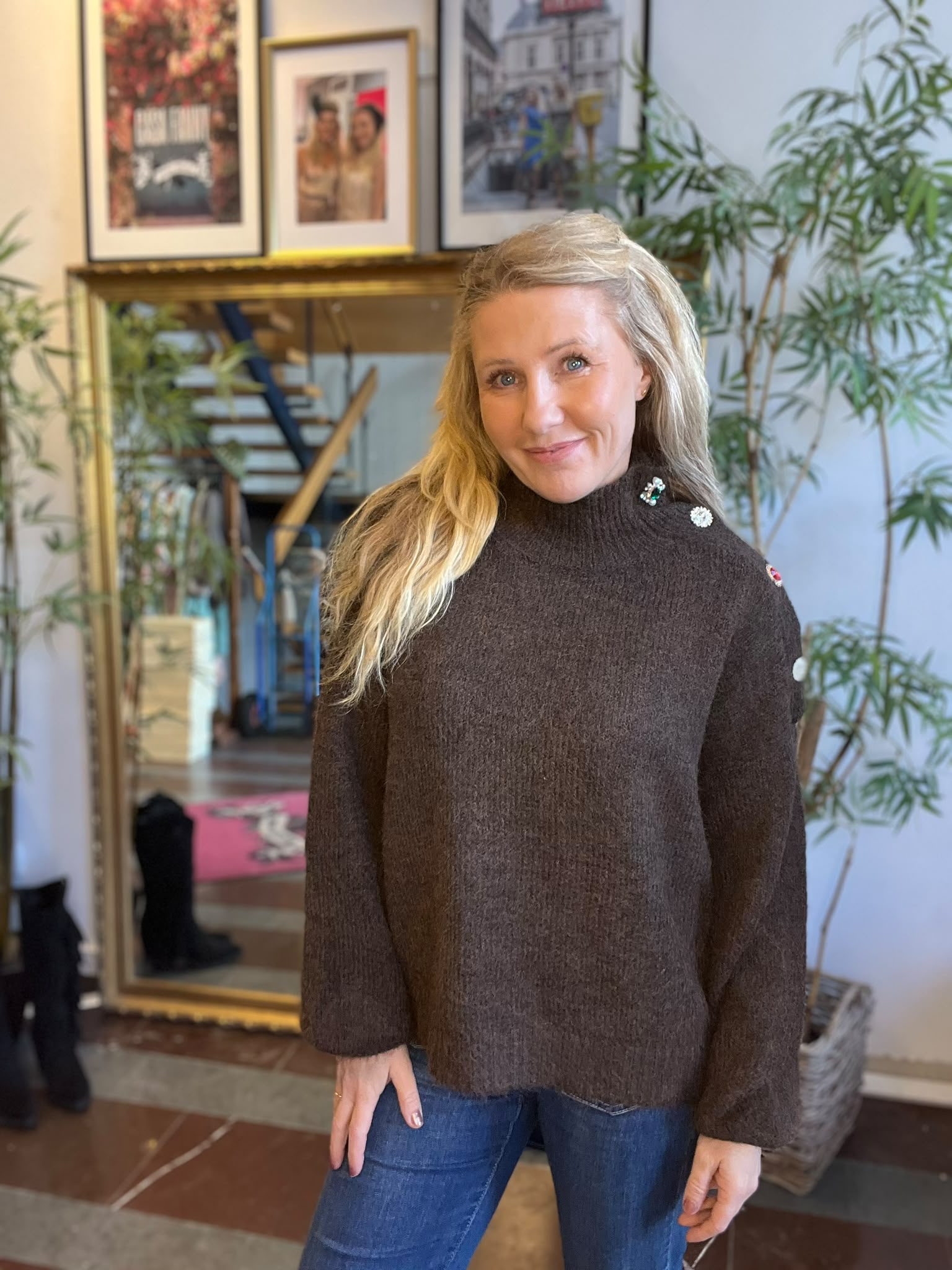 Blond kvinna står och ler mot kameran i ett hem, klädd i brun stickad tröja med dekorativa knappar vid axeln och jeans. Bakom henne syns spegel, inomhusväxter och inramade foton.