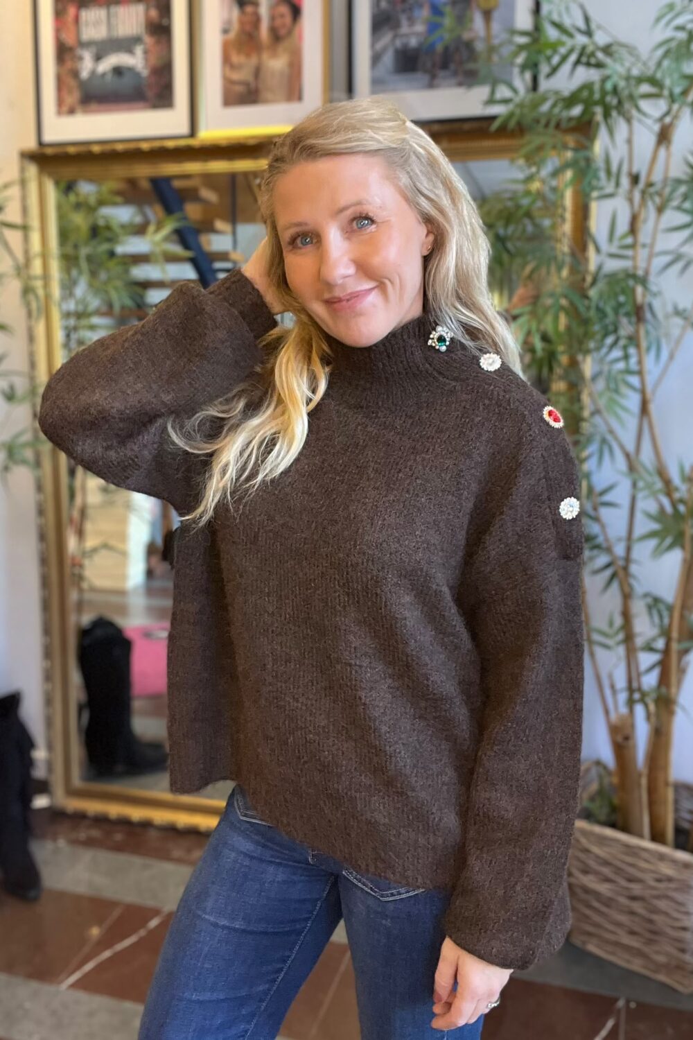 Blond kvinna poserar och ler mot kameran med ena handen bakom huvudet. Hon bär brun stickad tröja med dekorativa knappar på axeln och jeans, inomhus framför spegel, inramade foton och stora gröna växter.