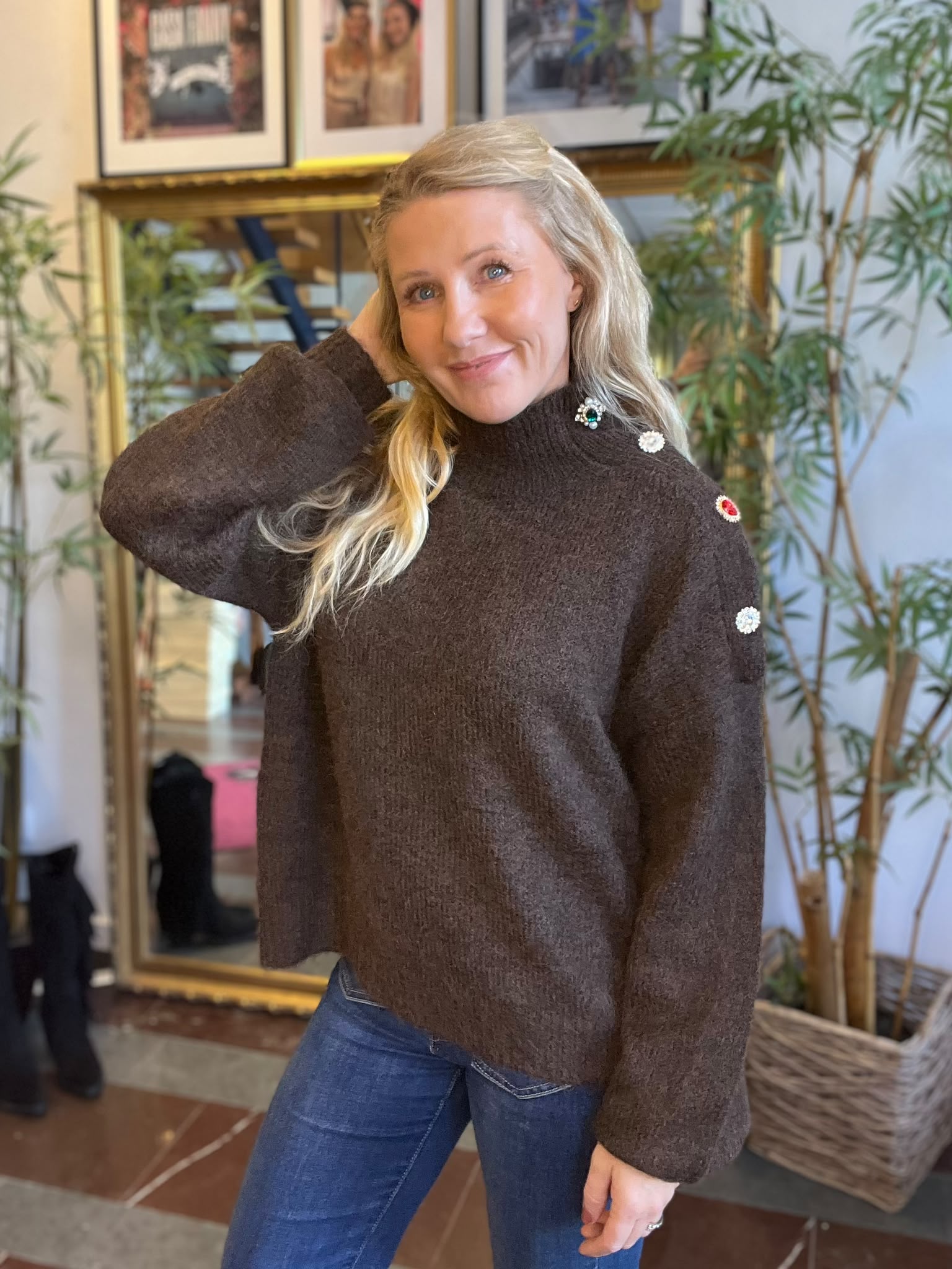 Blond kvinna poserar och ler mot kameran med ena handen bakom huvudet. Hon bär brun stickad tröja med dekorativa knappar på axeln och jeans, inomhus framför spegel, inramade foton och stora gröna växter.