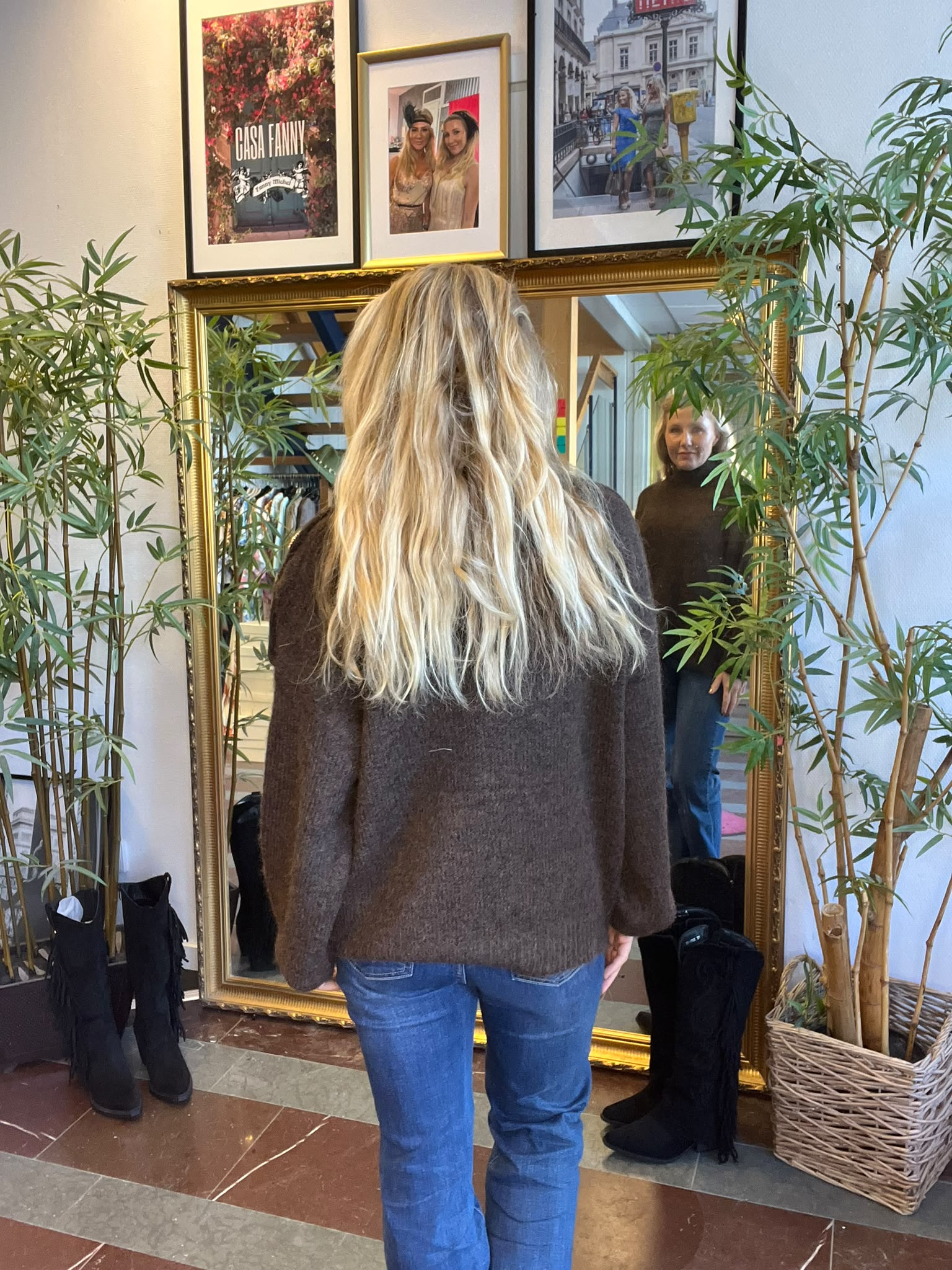 Kvinna med långt blont hår står vänd mot en stor guldramsspegel och ser sin spegelbild. Hon bär brun tröja och jeans; runt spegeln finns bambuplants. Ovanför hänger inramade foton, ett med texten “CASA FANNY”.