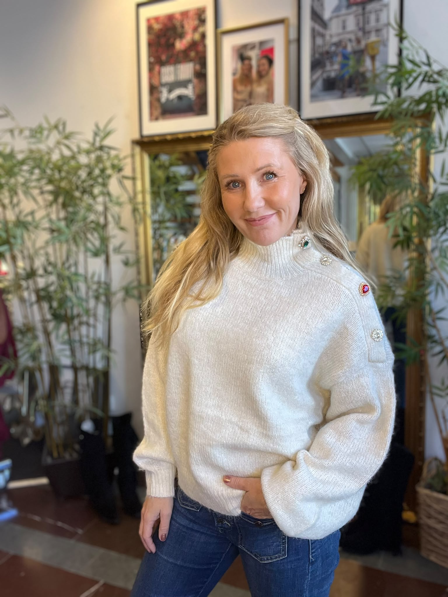 Blond kvinna poserar och ler mot kameran i ett ljust rum, klädd i vit polotröja med dekorativa knappar vid axeln och blå jeans. I bakgrunden syns gröna krukväxter och inramade foton.