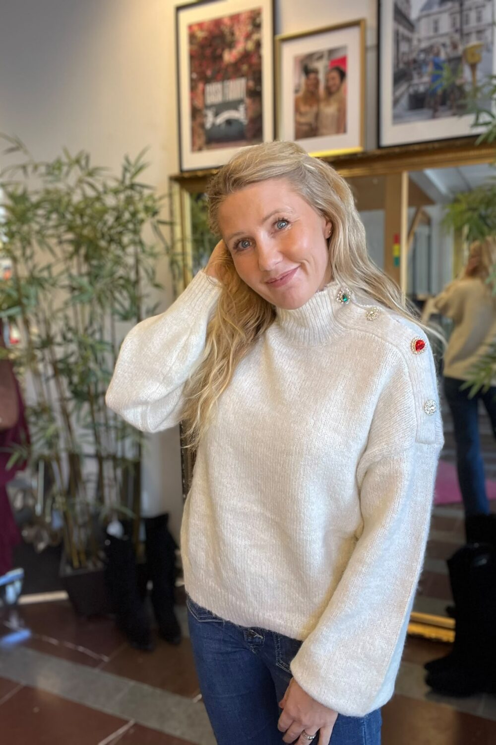 Blond kvinna i vit stickad polotröja med dekorativa knappar ler mot kameran och håller ena handen vid håret. Hon står inomhus i en butik eller salong, med växter, spegel och inramade foton i bakgrunden.
