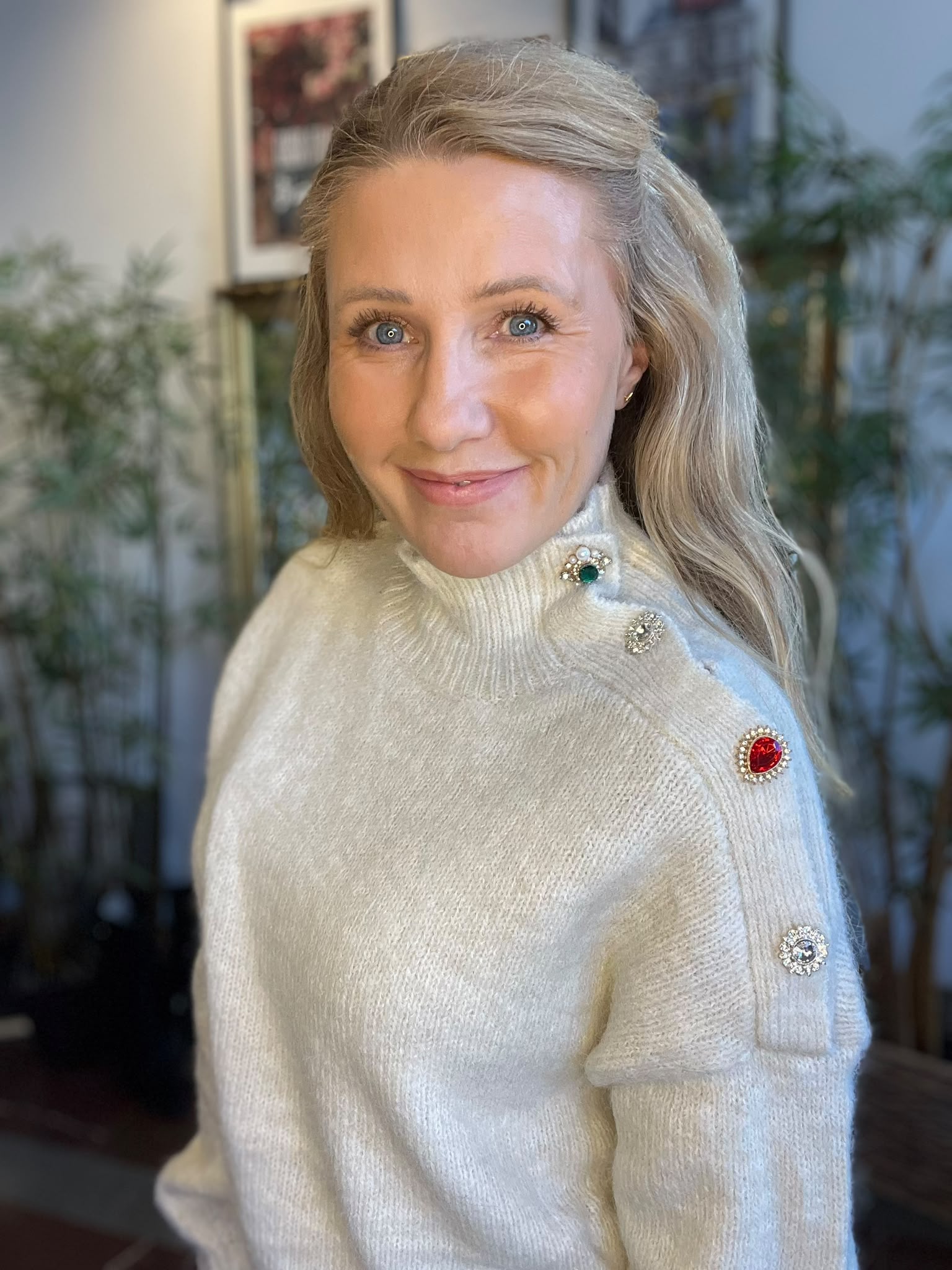 Leende kvinna med långt blont hår tittar mot kameran inomhus. Hon bär en vit, stickad polotröja med dekorativa knappnålar i rött, grönt och silver på axeln; suddig bakgrund med växter och tavla.