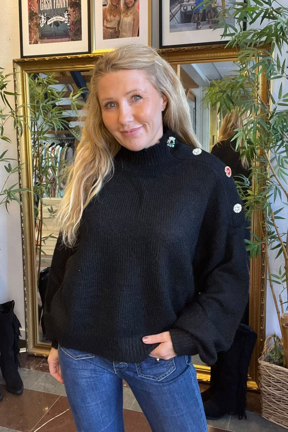 Blond kvinna står och ler mot kameran med ena handen i jeansfickan, klädd i svart stickad tröja med dekorativa knappar. Hon står framför en guldramsspegel, omgiven av gröna växter och inramade foton. Text: “GASA FANNY”.