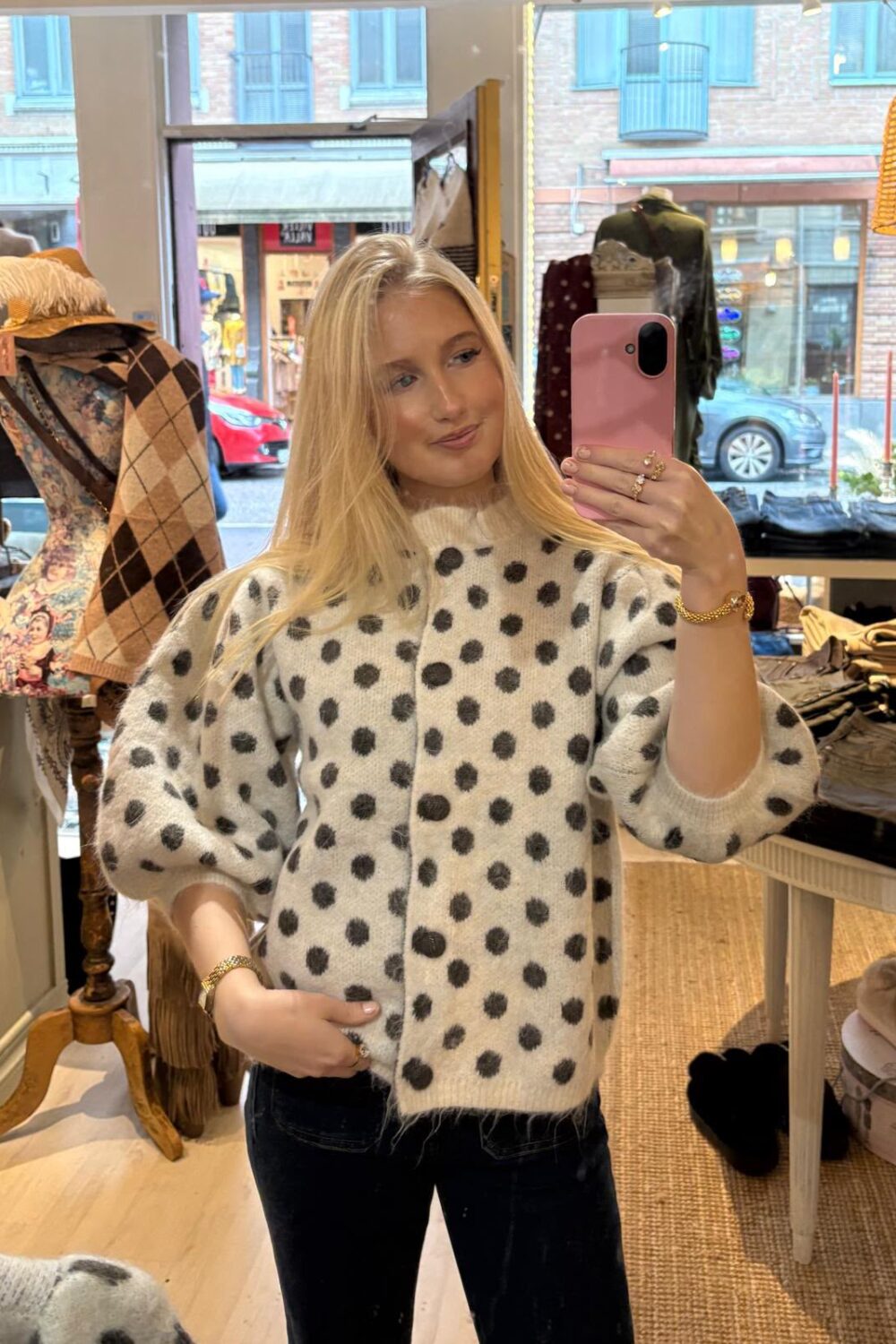 Blond person tar en spegelselfie med rosa mobil i en klädbutik och visar en vit kofta med stora svarta prickar. I bakgrunden syns skyltdocka med sjal och kläder på bord samt gata utanför fönstret.