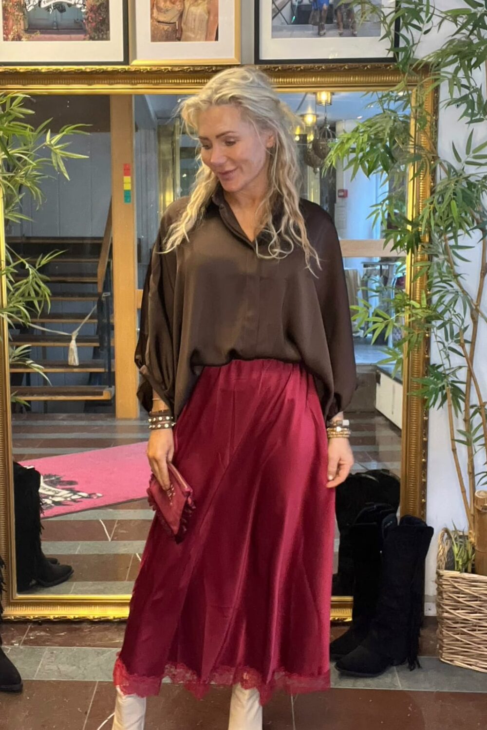 Kvinna med långt blont hår poserar framför en stor guldramsspegel, klädd i mörk blus och vinröd midikjol med spetskant, vita boots och vinröd kuvertväska. Bakom syns trappa, gröna växter och tavlor med texten ”CASA FANNY”.