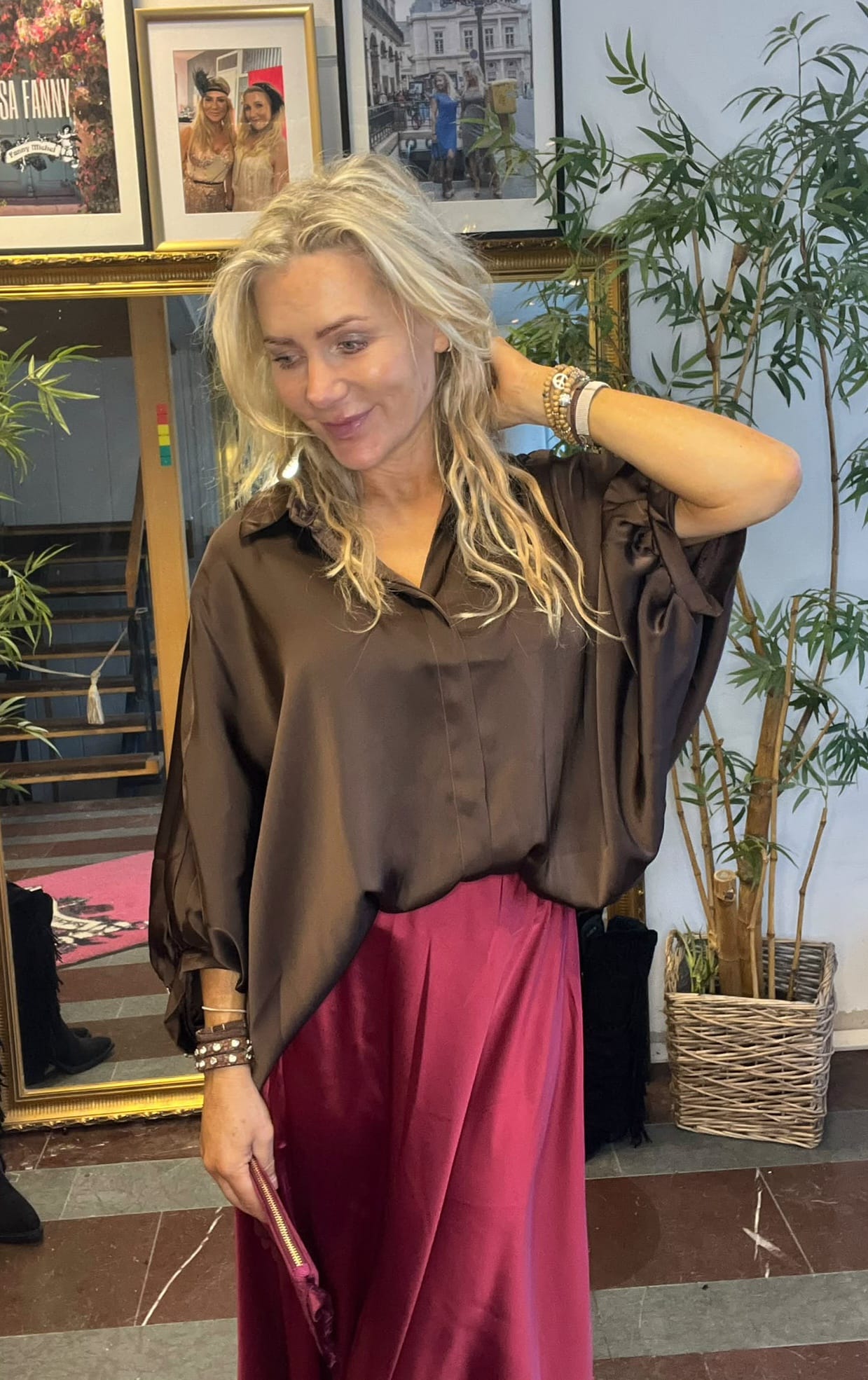 Blond kvinna står inomhus och ler lätt medan hon håller en hand bakom nacken och en rosa clutch i den andra. Hon bär brun blus och vinröd kjol, med inramade foton och krukväxter i bakgrunden.