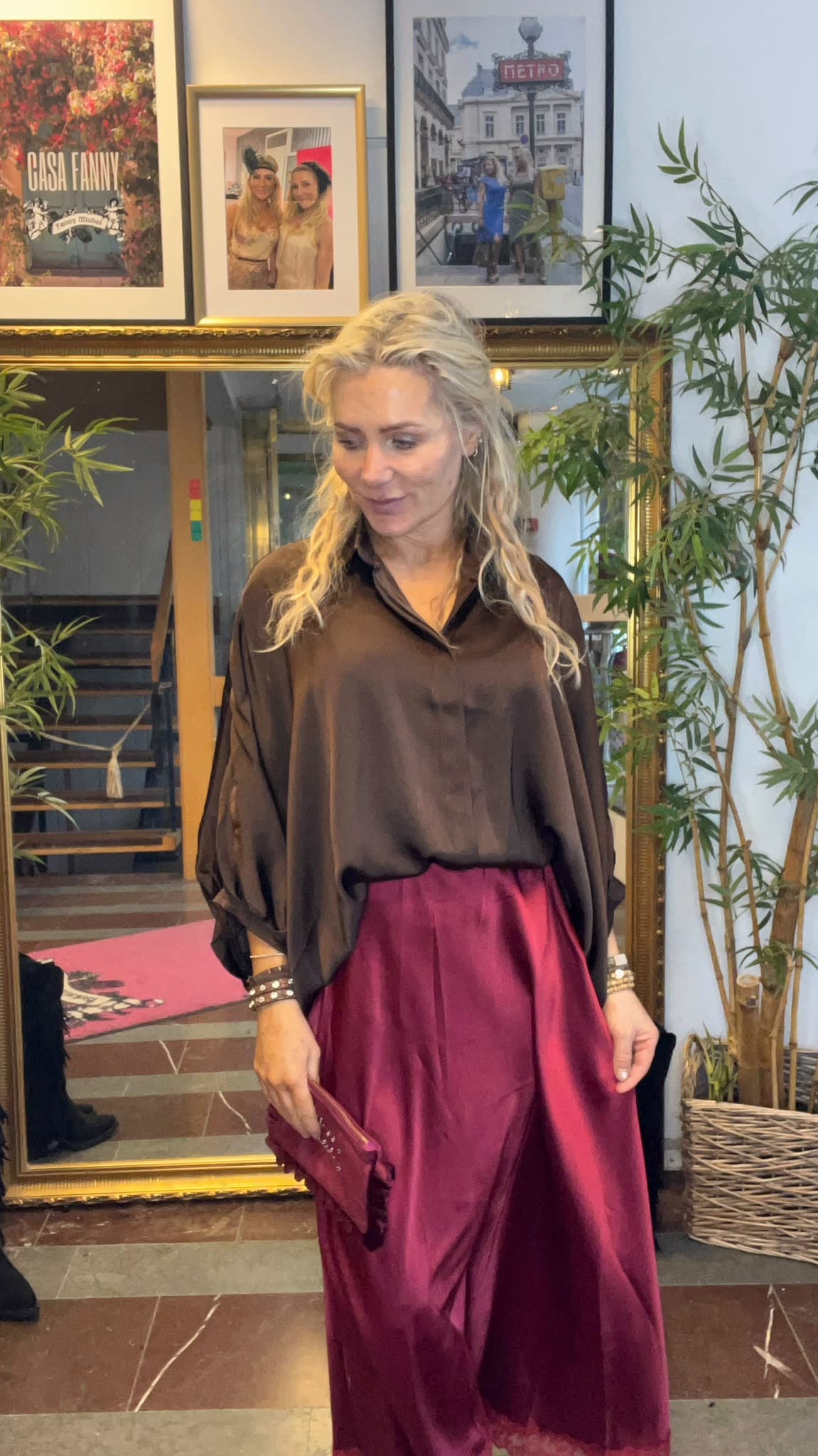 Blond kvinna står och ler svagt, klädd i svart blus och vinröd långkjol, med kuvertväska i handen. Hon står framför en stor spegel med trappa, växter och inramade foton; texten “CASA FANNY” syns.