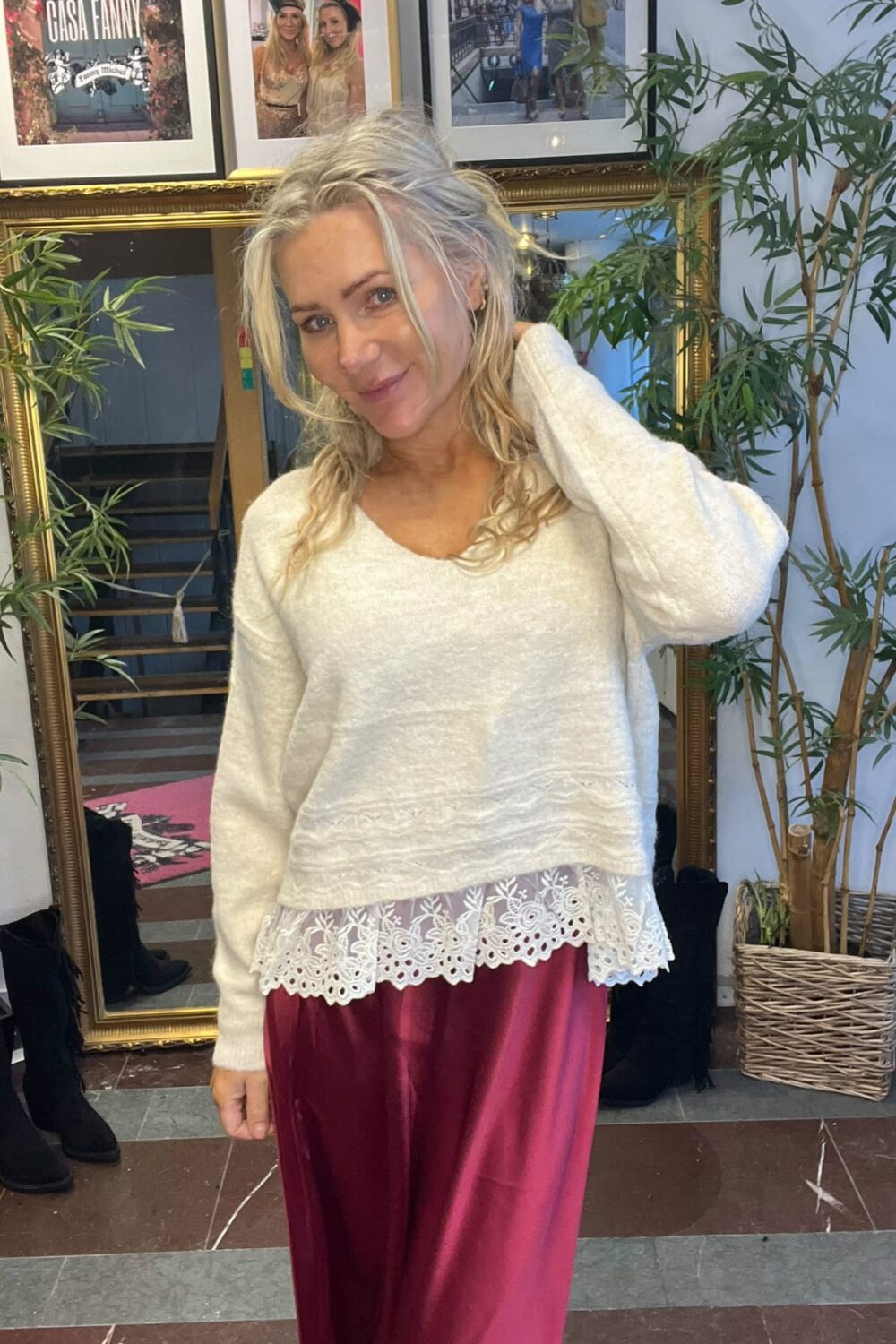 Leende kvinna med blont hår poserar och håller ena handen bakom nacken. Hon bär vit stickad tröja med spetskant och vinröd satinkjol, framför en guldramsspegel, gröna växter och inramade foton; text: “CASA FANNY”.