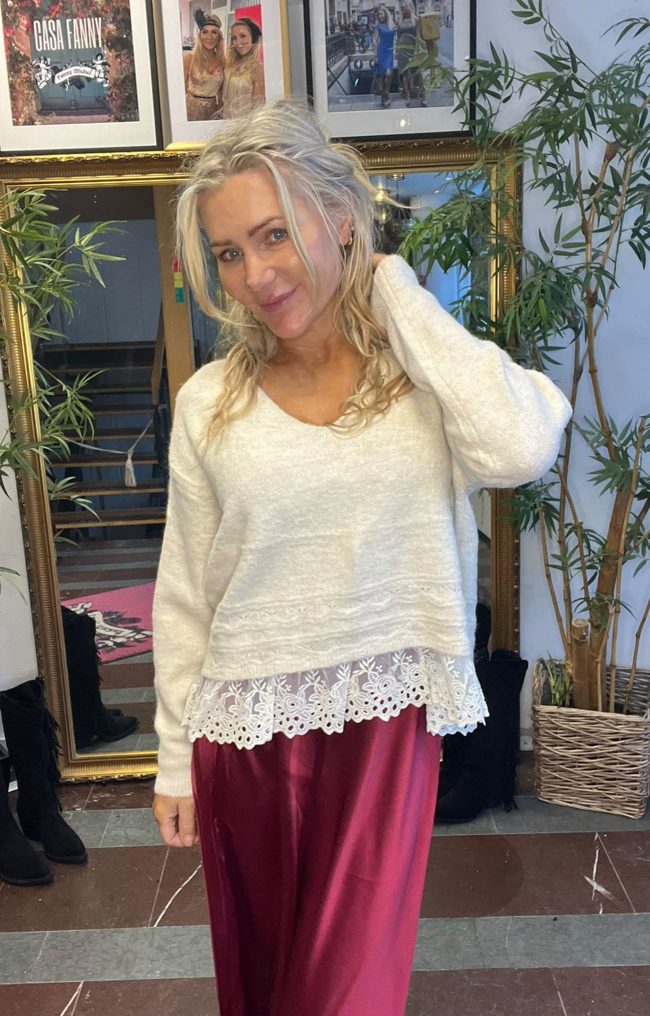 Leende kvinna med blont hår poserar och håller ena handen bakom nacken. Hon bär vit stickad tröja med spetskant och vinröd satinkjol, framför en guldramsspegel, gröna växter och inramade foton; text: “CASA FANNY”.