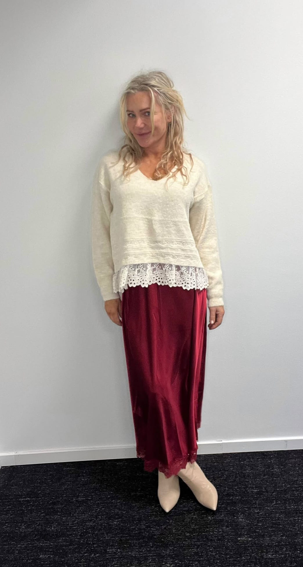 Blond kvinna står och poserar mot en ljus vägg, med lätt leende och blicken åt sidan. Hon bär en ljus stickad tröja med spetskant, vinröd lång kjol och beige spetsiga skor.