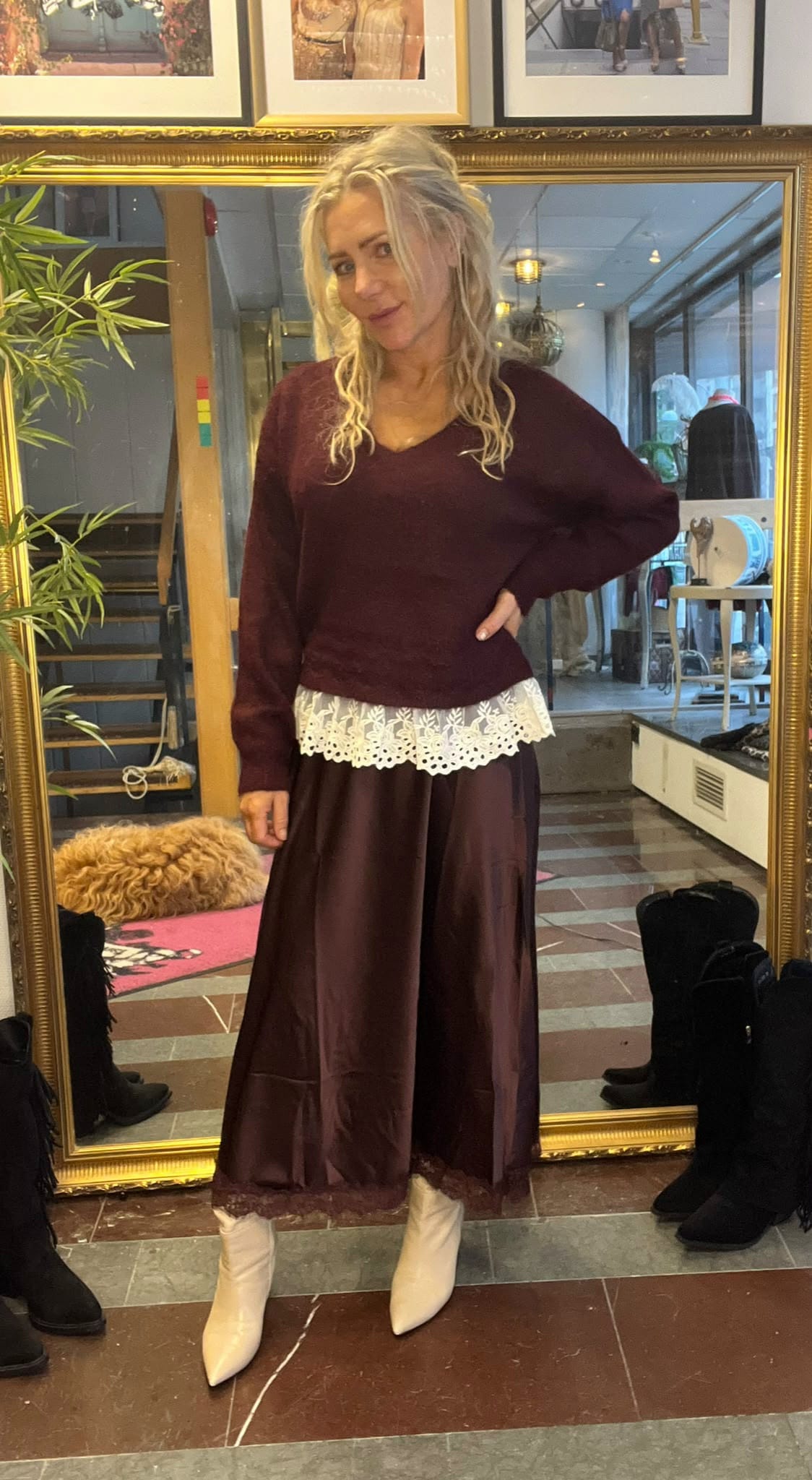 Kvinna med blont, lockigt hår poserar med handen på höften framför en stor guldramsspegel. Hon bär vinröd tröja, brun lång kjol med vit spetskant och vita boots; butiksmiljö med skor och inredning syns runtom.