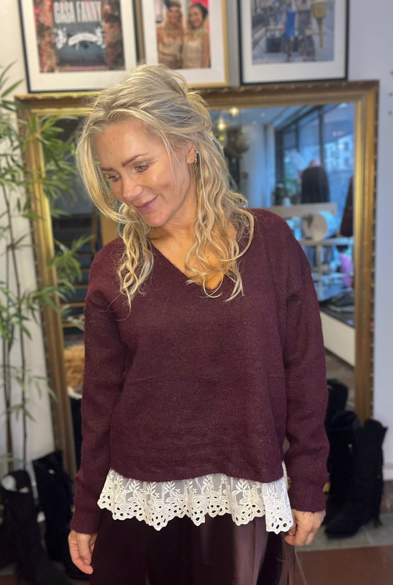Blond kvinna står inomhus och ler medan hon tittar ned åt sidan. Hon bär vinröd stickad tröja med vit spetskant över en mörk kjol, framför en stor guldramsspegel och inramade foton på väggen.
