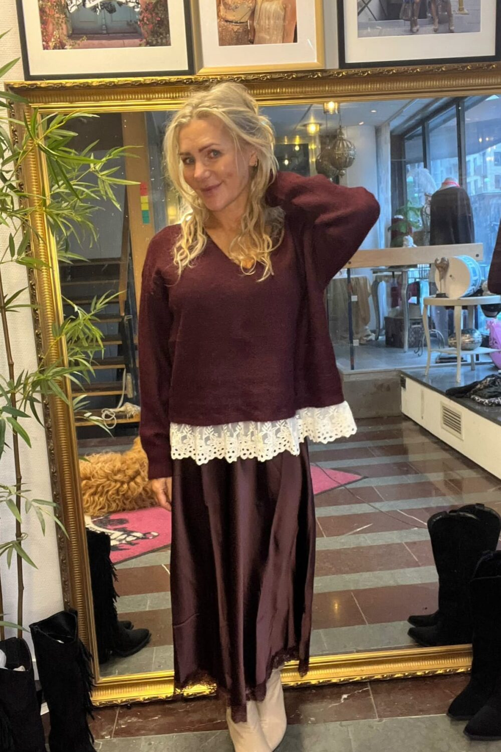 Blond kvinna står och poserar med ena handen bakom nacken framför en stor guldramsspegel i en butik. Hon bär vinröd tröja med vit spetskant, lång brun kjol och vita stövlar; stövlar och inramade foton syns runtom.
