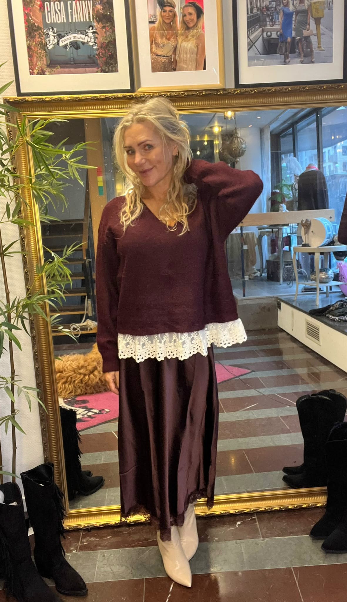 Blond kvinna poserar framför en stor guldramad spegel, med handen bakom huvudet och ett leende. Hon bär vinröd tröja med vit spetskant, lång mörk kjol och vita boots, i en butiksmiljö med stövlar och växter. Text på tavla: “CASA FANNY”.