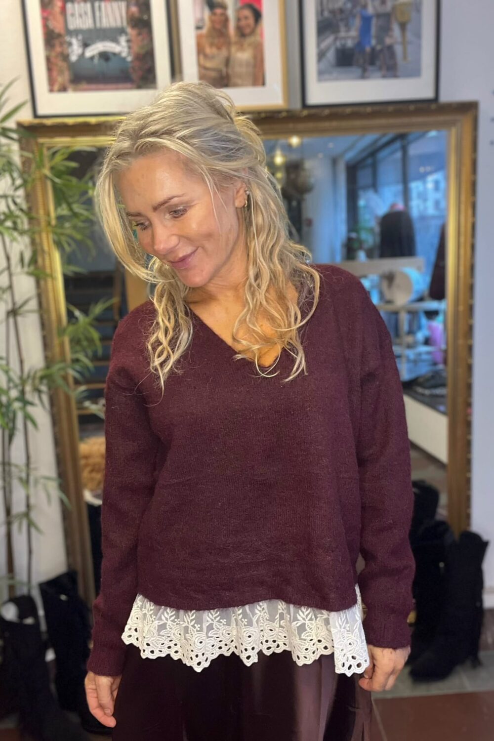 Leende blond kvinna står inomhus och tittar ner åt sidan, klädd i vinröd stickad tröja med vit spetskant över en mörk kjol. Bakom henne syns stor guldramsspegel, inramade foton och en grön växt.