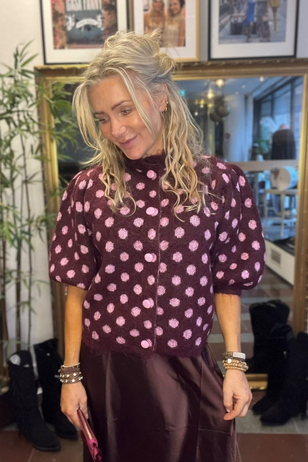 Blond kvinna står och ler nedåt, klädd i vinröd prickig kofta med rosa knappar och mörk satinkjol, med armband och en liten väska i handen. Bakom henne syns en stor spegel och inramade foton.