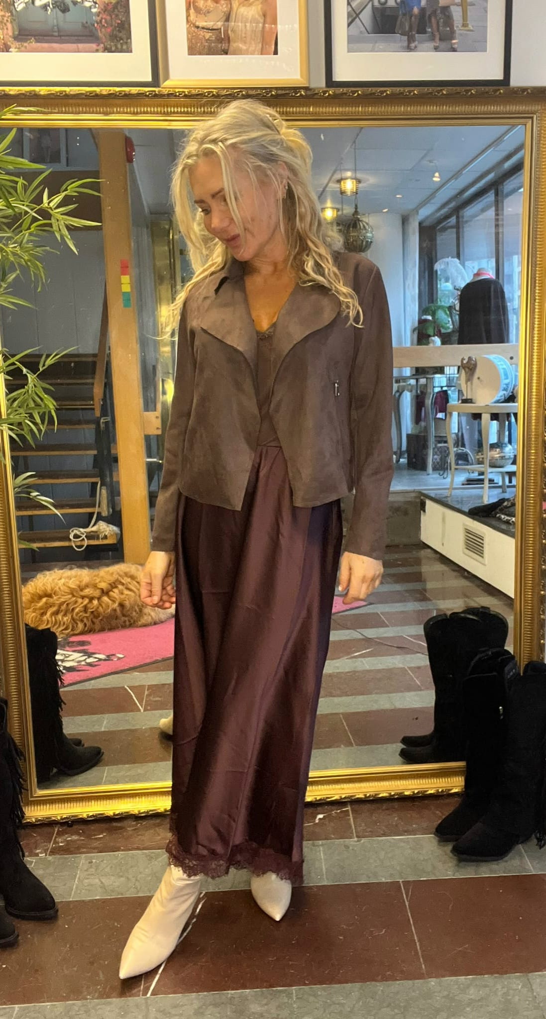 Blond kvinna står framför en stor guldramsspegel och tittar ned med ett leende, klädd i vinröd satinklänning med spetskant och brun kort jacka samt vita spetsiga stövlar. Bakom syns butiksmiljö med trappa och kläder.