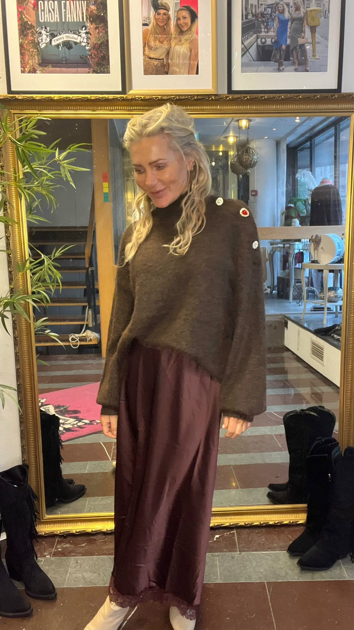Blond kvinna står och provar en outfit framför en stor guldramsspegel i en butik, klädd i brun stickad tröja med dekorativa knappar och vinröd satinkjol. På väggen ovanför syns inramade foton och texten “CASA FANNY”.