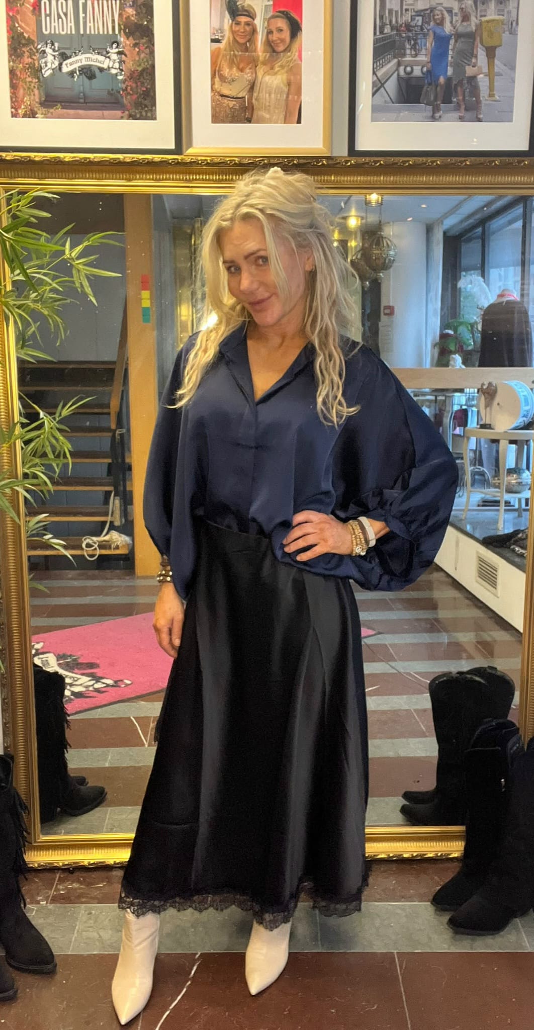Blond kvinna poserar med handen på höften framför en stor guldramsspegel, klädd i mörkblå blus och svart kjol med vita boots. I bakgrunden syns trappa, växter och inramade foton med texten “CASA FANNY”.