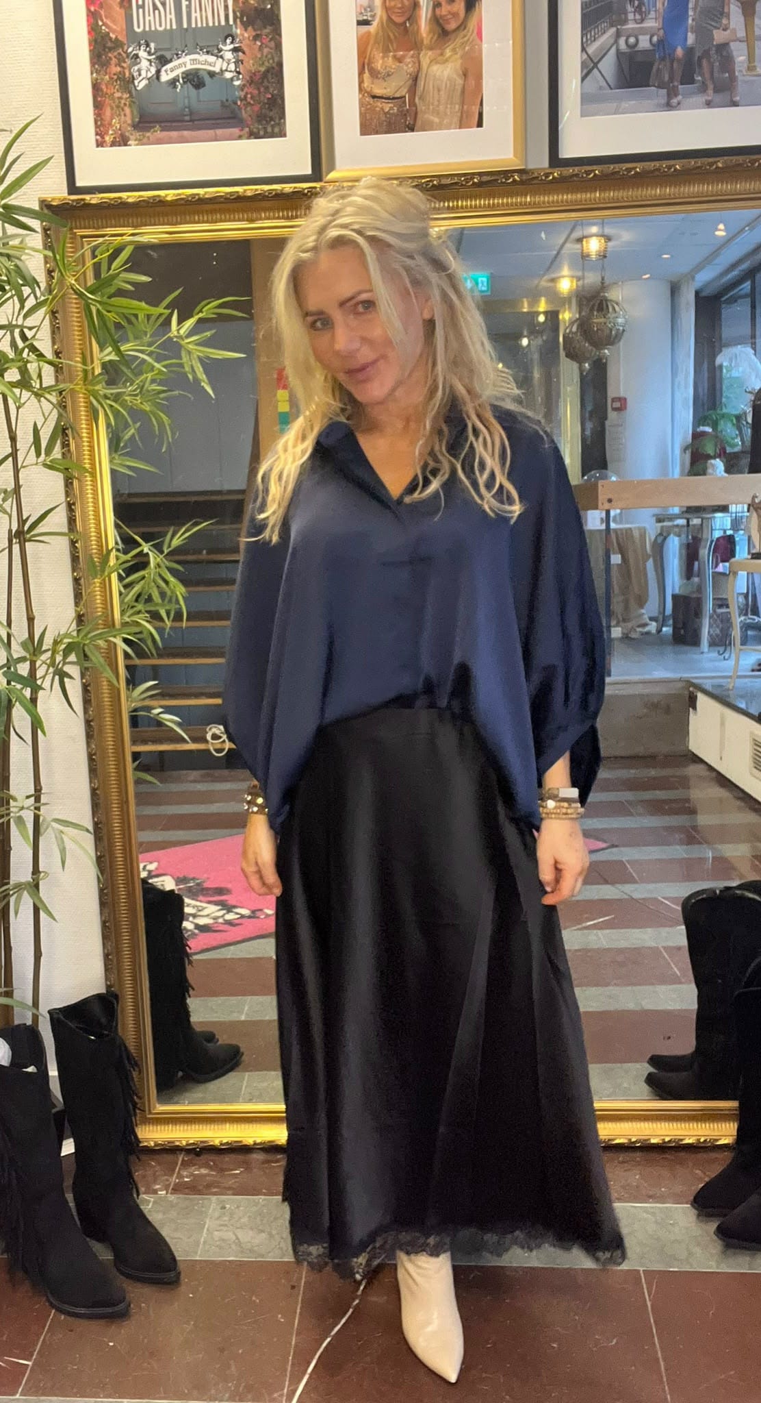 Kvinna med långt blont hår poserar och ler svagt framför en stor guldramsspegel, klädd i mörkblå blus och svart satinkjol med vita spetsiga boots. I bakgrunden syns trappa, växt och tavlor med texten ”CASA FANNY” och ”Fanny Michel”.