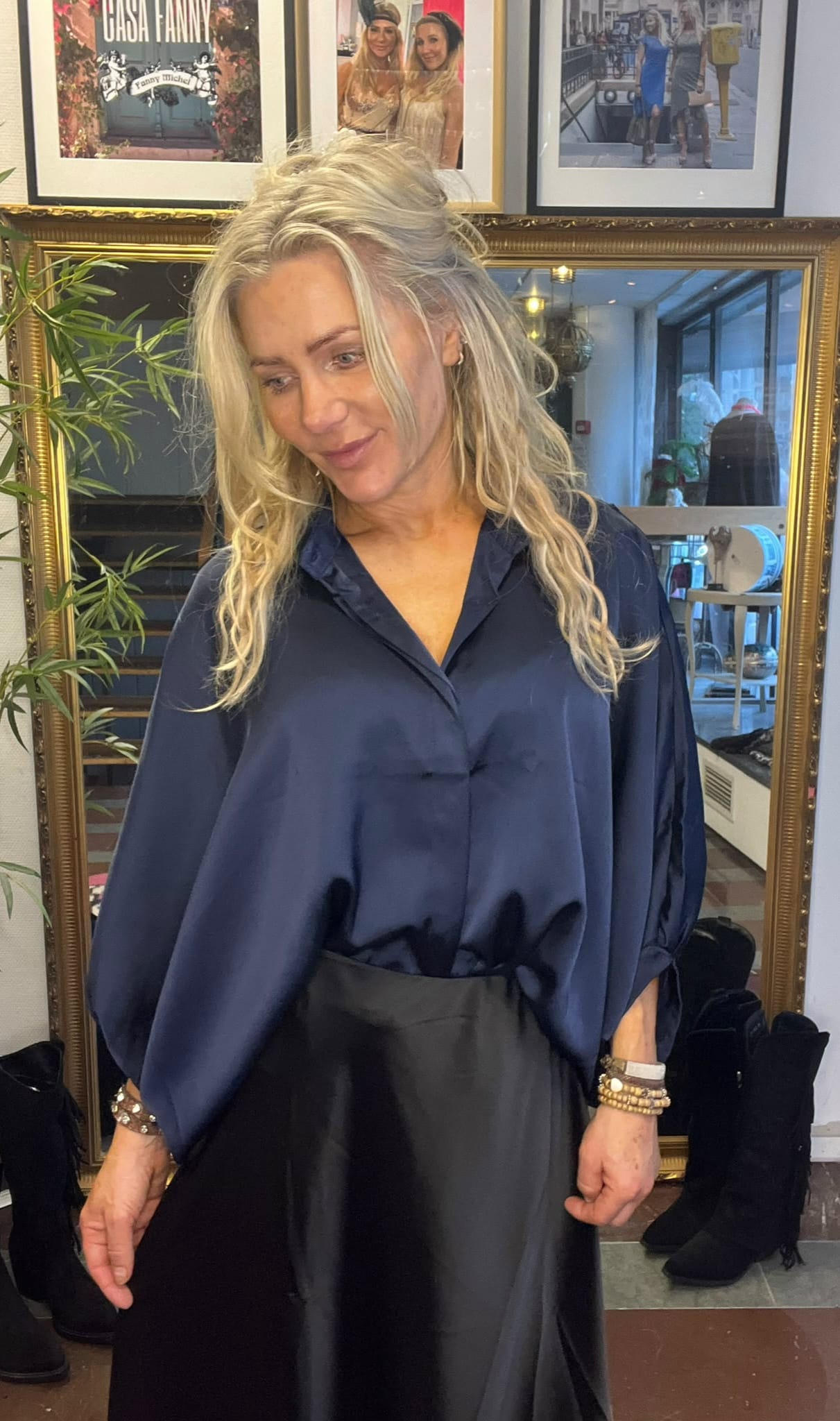 Blond kvinna står och tittar nedåt med ett svagt leende, klädd i mörkblå blus och svart kjol. Hon poserar framför en stor guldramsspegel i en butik, med växt och inramade foton ovanför. Text: “CASA FANNY”.