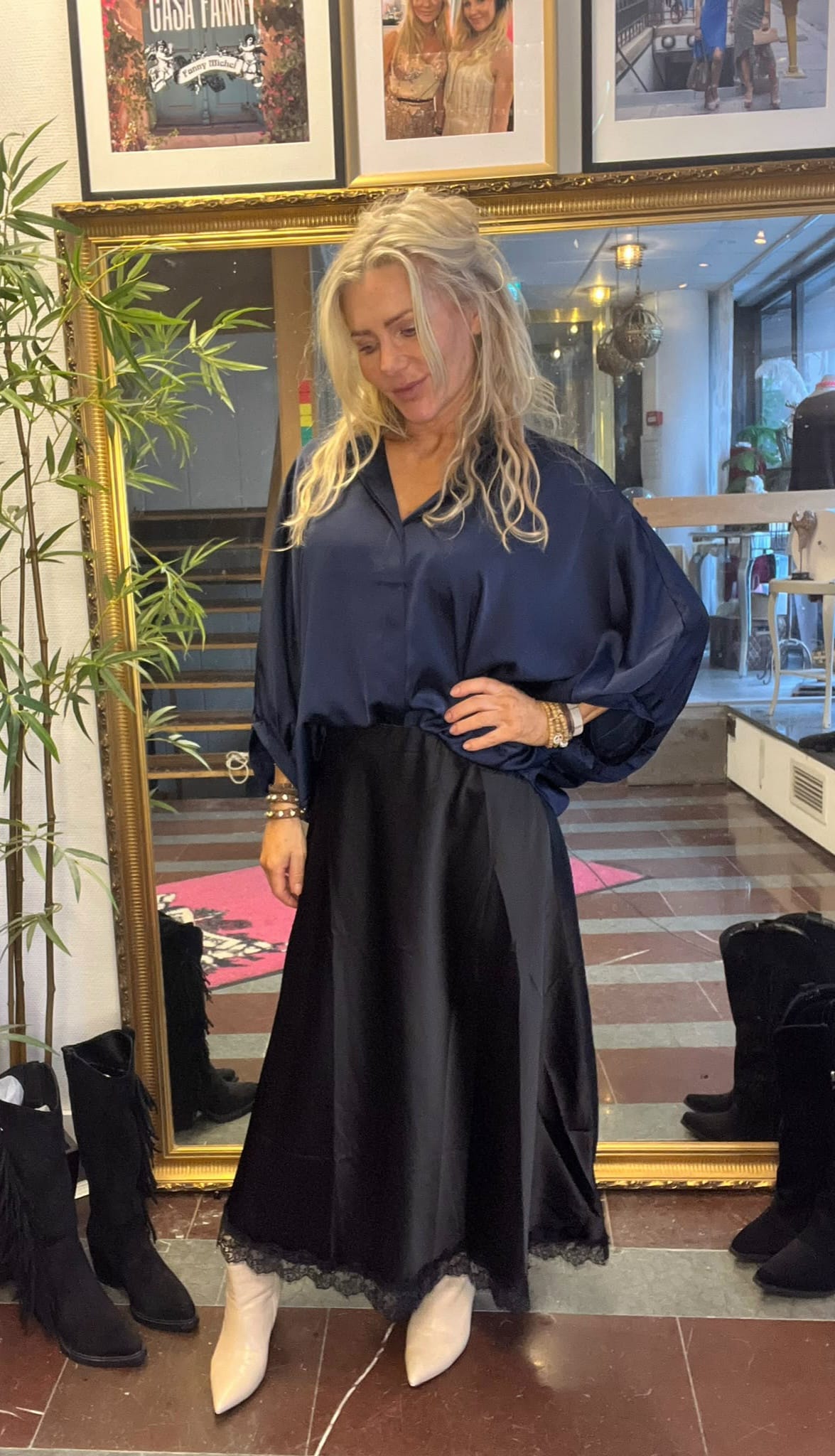 Blond kvinna poserar med handen på höften framför en stor guldramsspegel och tittar nedåt. Hon bär mörkblå blus, lång svart kjol med spetskant och vita boots; i bakgrunden syns butikslokal, växt och inramade foton.