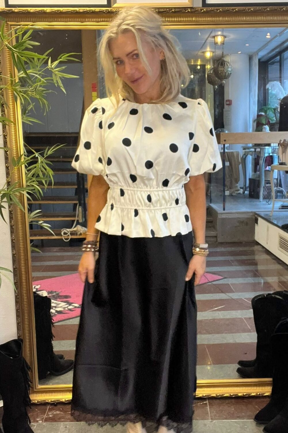 Blond kvinna står och poserar framför en stor guldramig spegel i en butiksmiljö, iklädd vit blus med svarta prickar och lång svart kjol med vita spetsiga skor. Ovanför syns inramade foton. Text: “CHA CHA FANNY”, “Fanny Michel”.