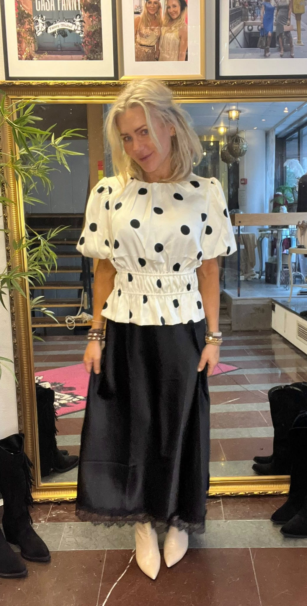 Blond kvinna står och poserar framför en stor guldramig spegel i en butiksmiljö, iklädd vit blus med svarta prickar och lång svart kjol med vita spetsiga skor. Ovanför syns inramade foton. Text: “CHA CHA FANNY”, “Fanny Michel”.