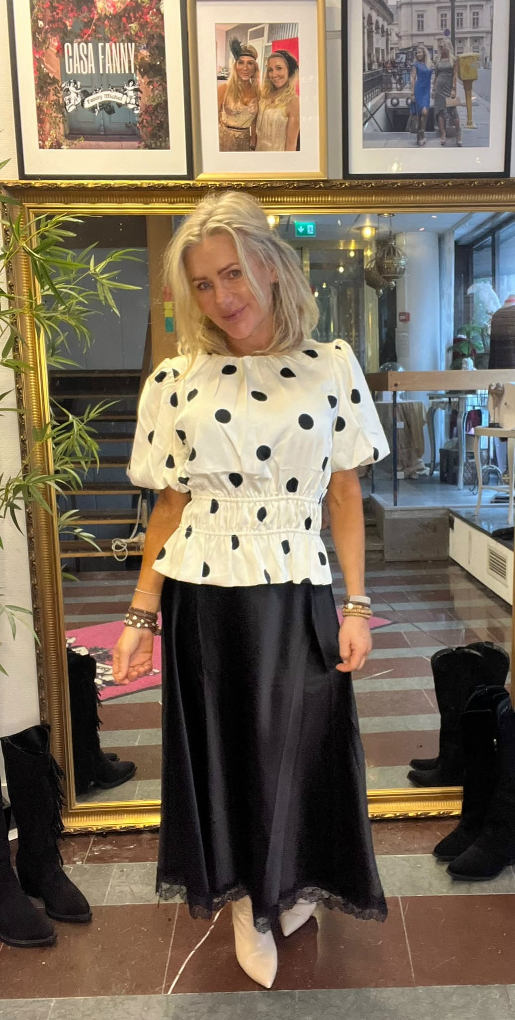 Blond kvinna poserar och ler framför en stor guldramad spegel, klädd i vit prickig blus, svart satinkjol med spetskant och vita stövlar. I bakgrunden syns inramade foton och texten “CASA FANNY”.