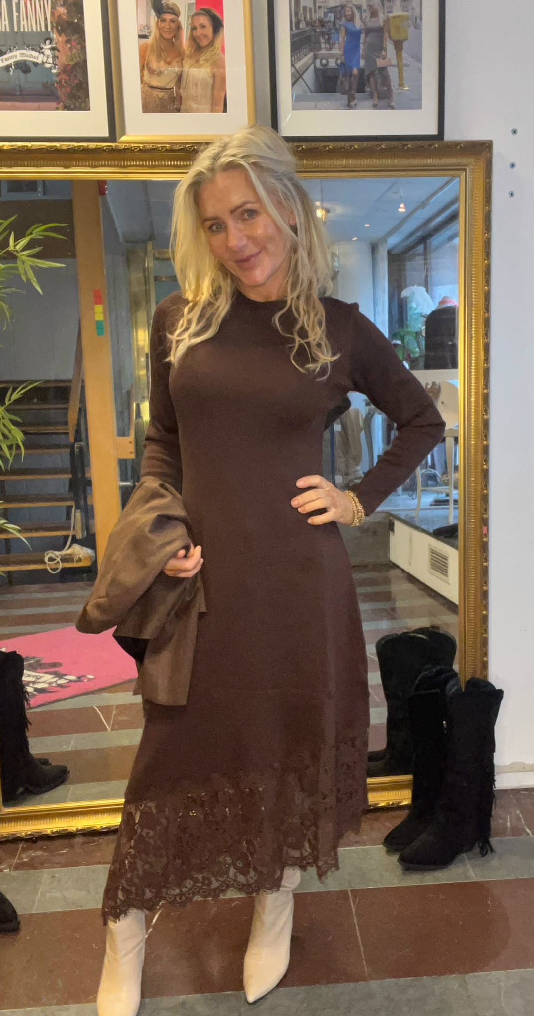 Blond kvinna poserar framför en stor guldramsspegel, med handen på höften och en jacka i andra handen. Hon bär brun långärmad klänning med spetskant och vita boots, i en butikslik miljö med foton på väggen.