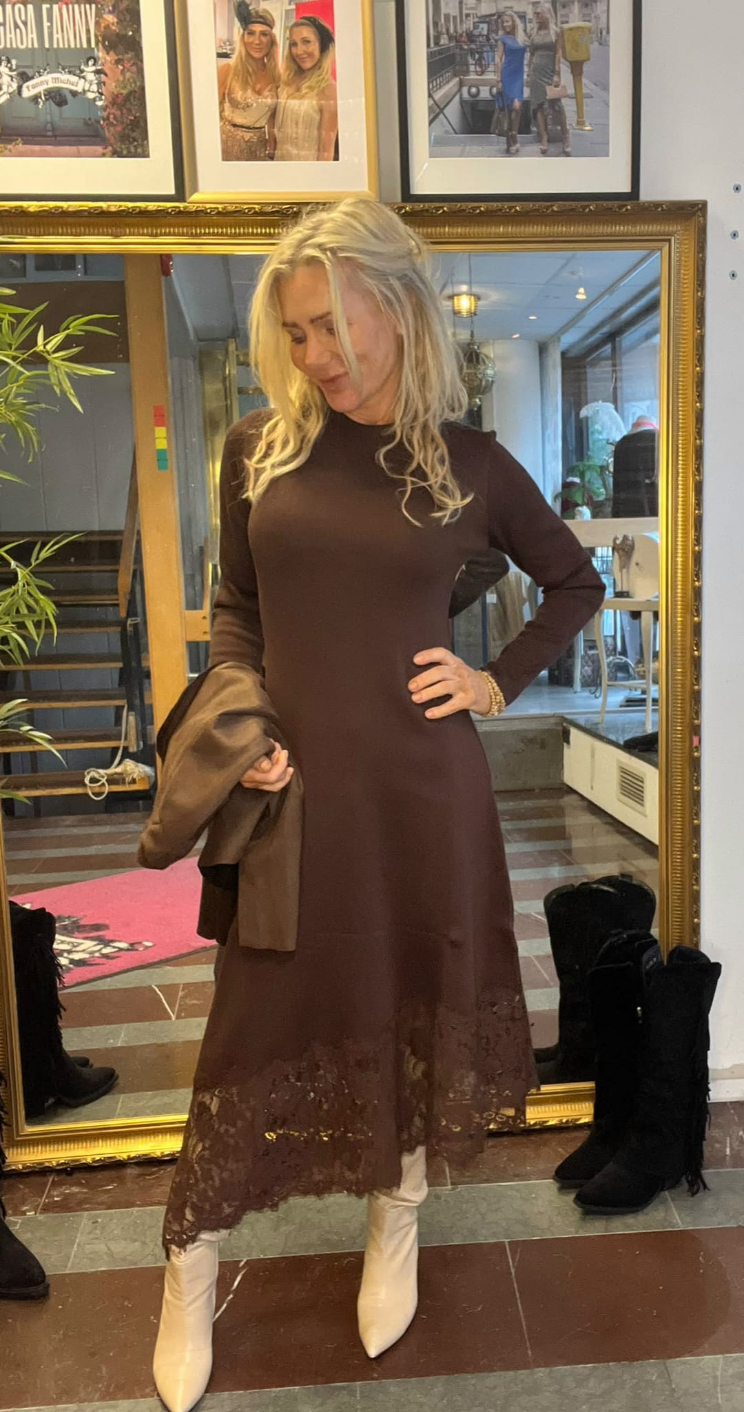 Blond kvinna posar framför en stor guldramsspegel, med handen på höften och en brun kappa över armen. Hon bär en långärmad brun klänning med spetskant och vita stövlar, i en butiksmiljö med trappa och fotografier på väggen.