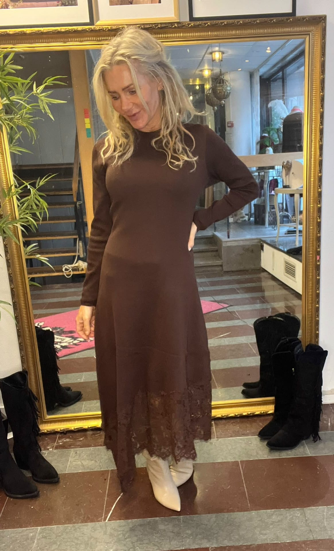 Kvinna provar en brun, långärmad midiklänning med spetskant och står framför en stor guldramad spegel, med handen på höften och blicken nedåt. Hon bär vita boots; i bakgrunden syns butikslokal och flera svarta stövlar.