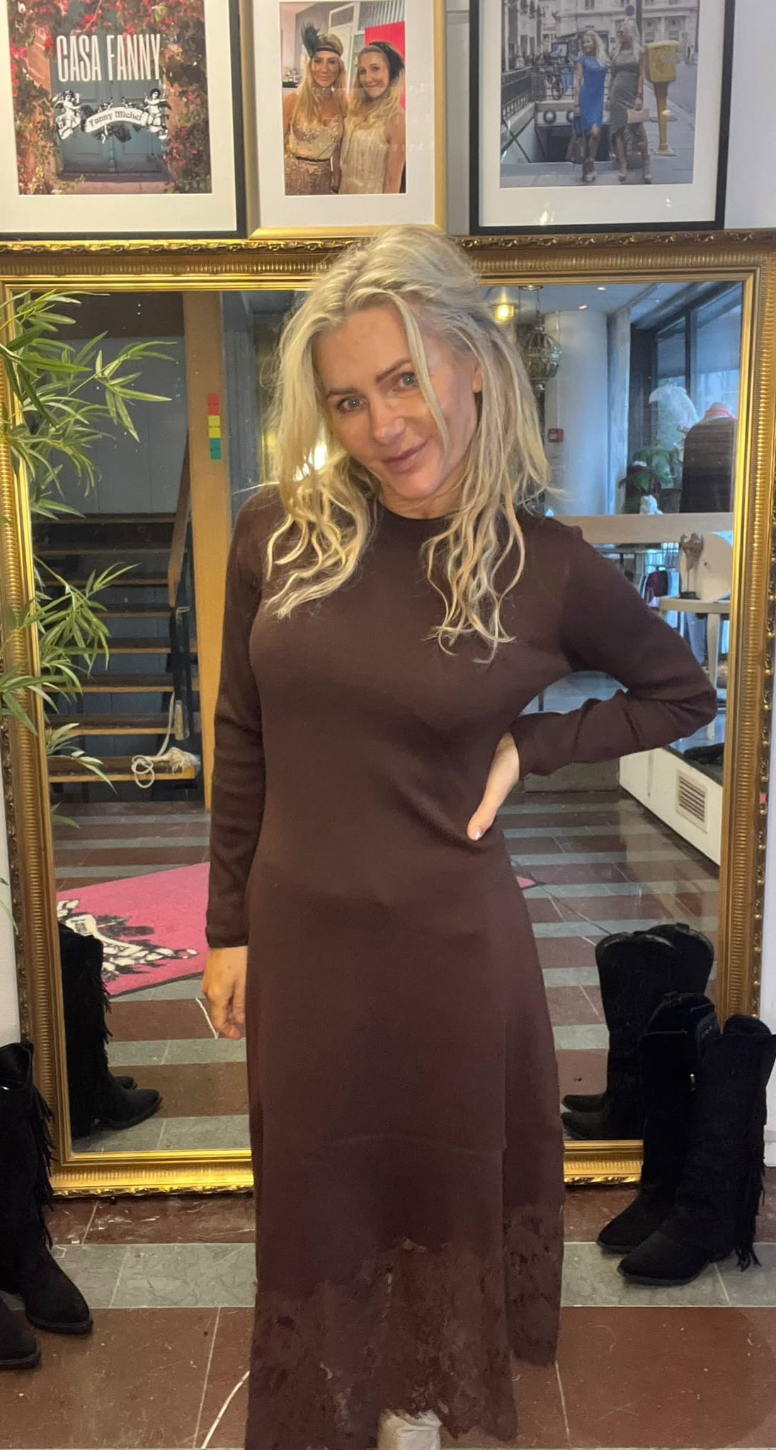Kvinna med långt blont hår poserar leende med handen på höften framför en stor guldramad spegel i en butik eller ateljé. Hon bär en lång brun klänning; bakom syns stövlar och inramade foton, bland annat texten “CASA FANNY”.