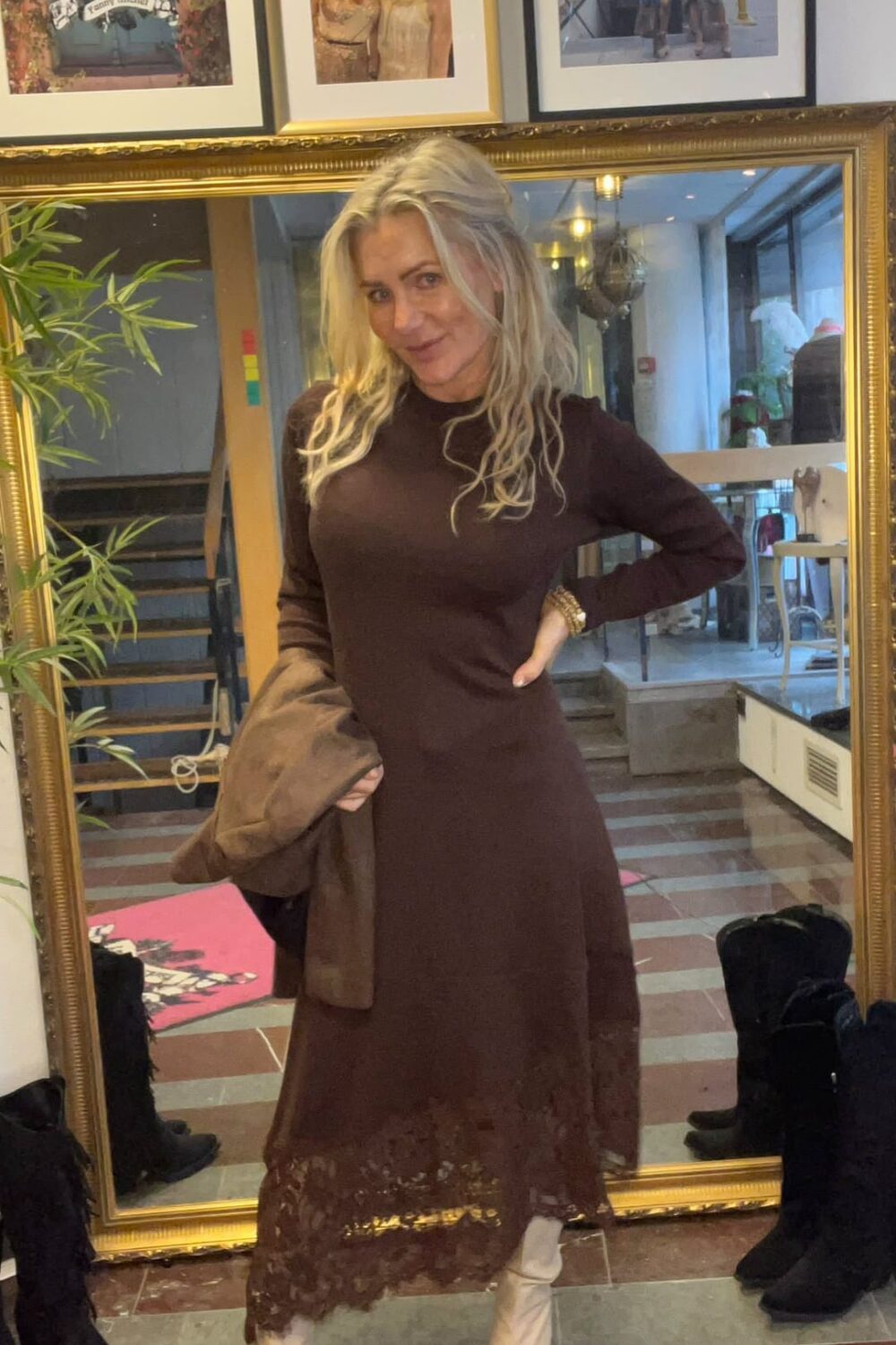 Blond kvinna poserar framför en stor guldramsspegel, med ena handen i sidan och en kappa i den andra. Hon bär en lång brun klänning med spetskant och vita stövlar, i en hall med trappa och inramade foton. Text: “CASA FANNY”.