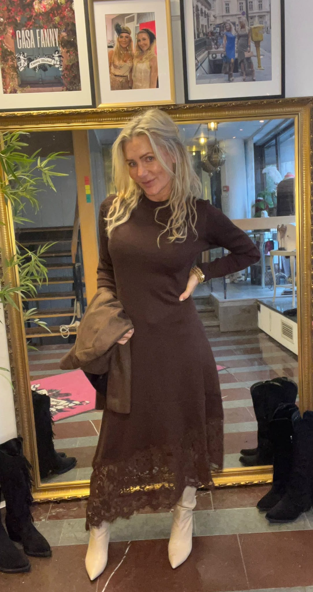 Blond kvinna poserar framför en stor guldramsspegel, med ena handen i sidan och en kappa i den andra. Hon bär en lång brun klänning med spetskant och vita stövlar, i en hall med trappa och inramade foton. Text: “CASA FANNY”.