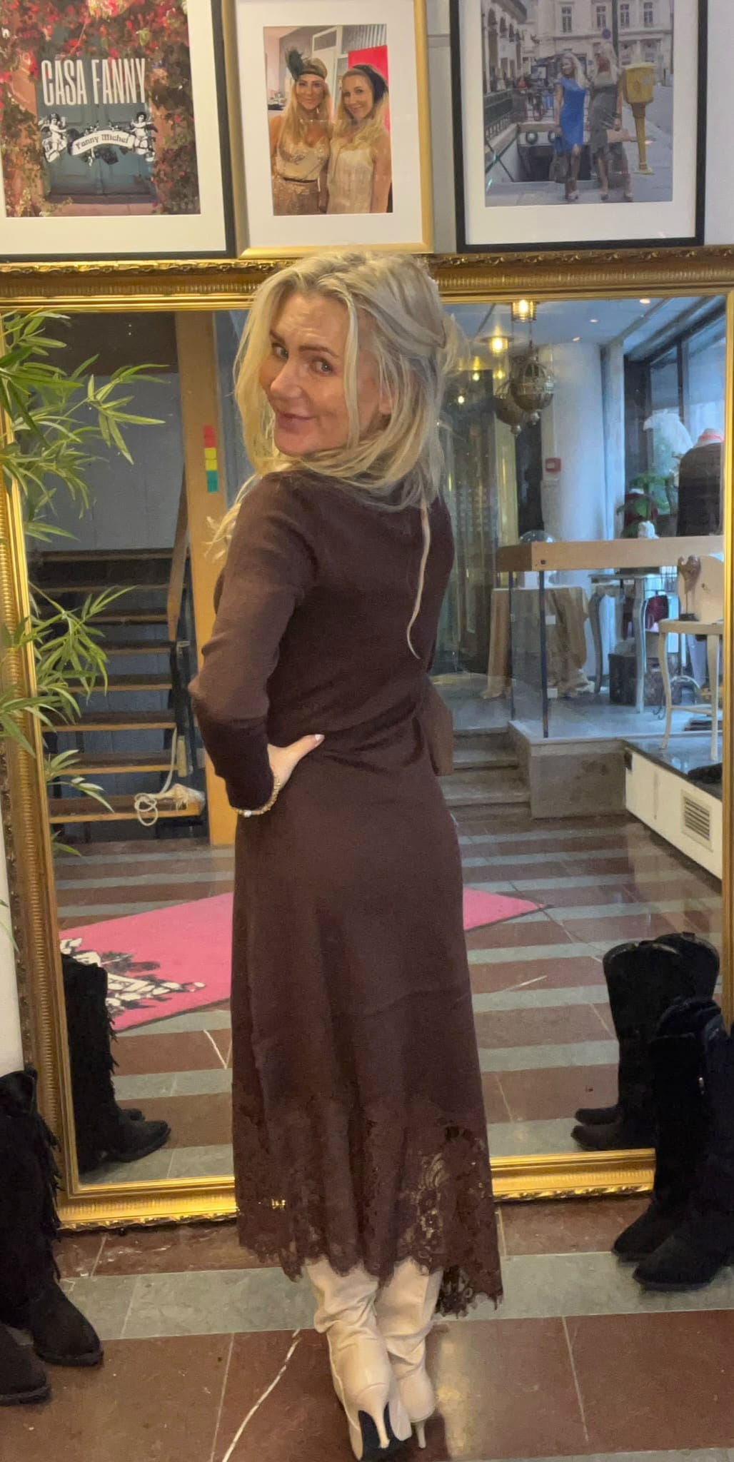 Kvinna med långt blont hår poserar med handen på höften och tittar över axeln mot kameran, iförd brun klänning med spetskant och vita stövlar, framför en stor spegel i en butikssalong med inramade foton ovanför. Text: ”CASA FANNY”, ”Fanny Unched”.