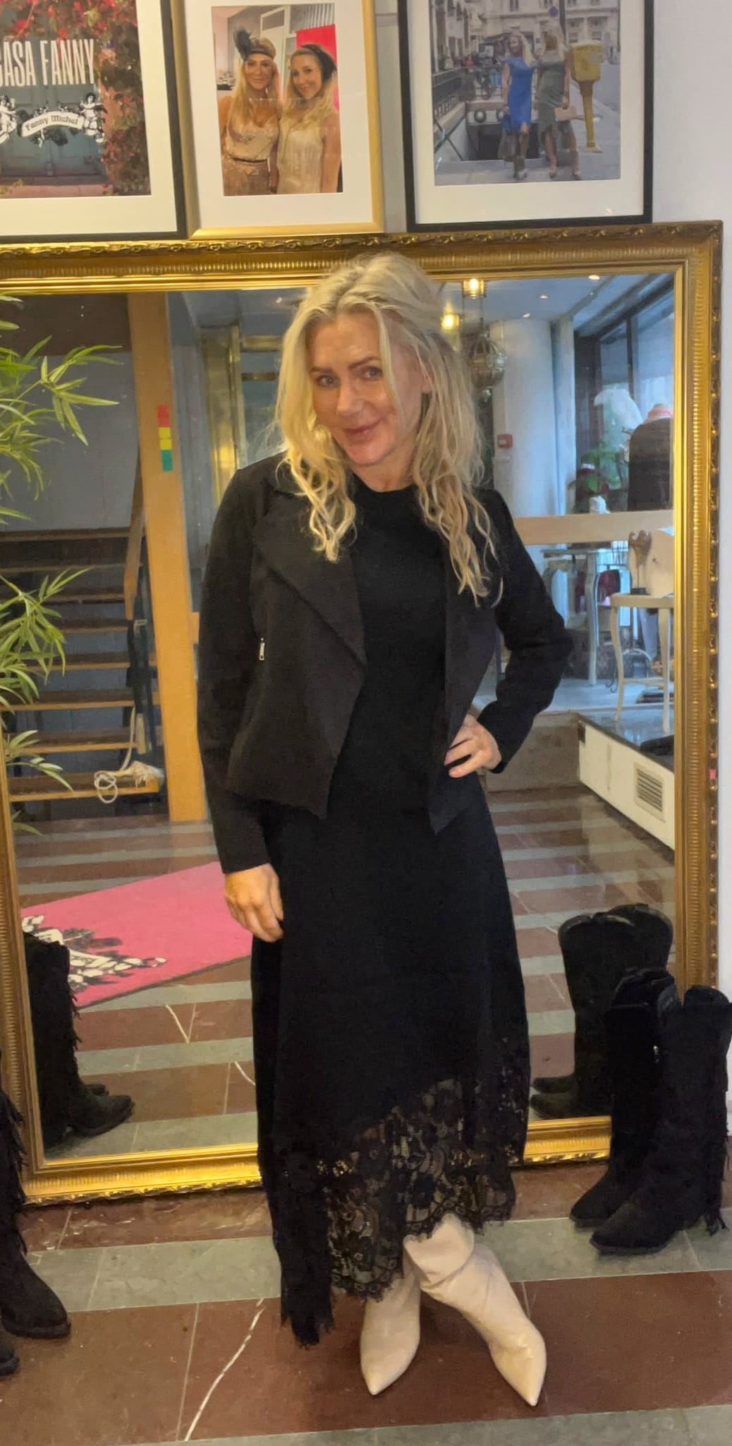 Blond kvinna poserar med handen på höften framför en stor guldramsspegel, klädd i svart klänning med spetskant och svart jacka samt vita stövlar. I bakgrunden syns trappa, växter och inramade foton på väggen.