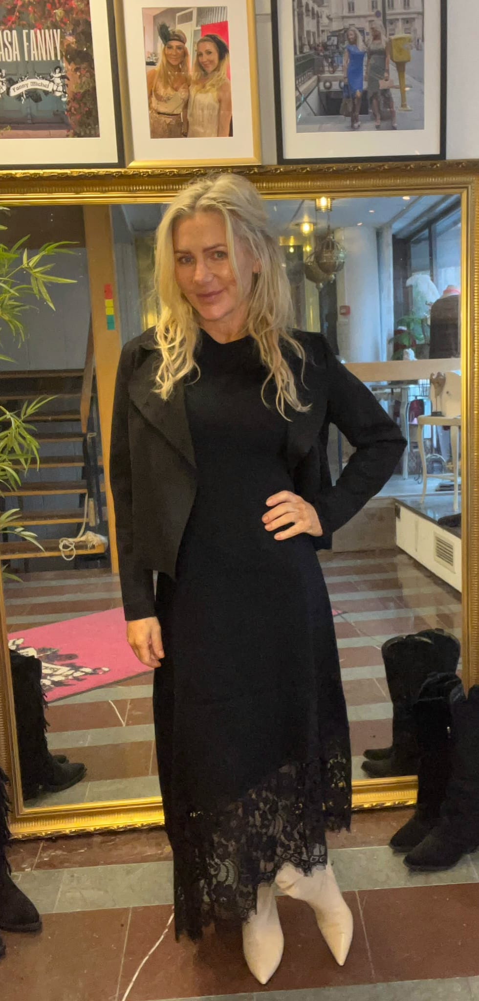 Blond kvinna står och poserar med handen på höften framför en stor guldramsspegel, klädd i svart långklänning med spetskant och vitspetsiga stövlar. I bakgrunden syns trappa, växter och inramade foton på väggen. Text på bild: “ASH FANNY”.